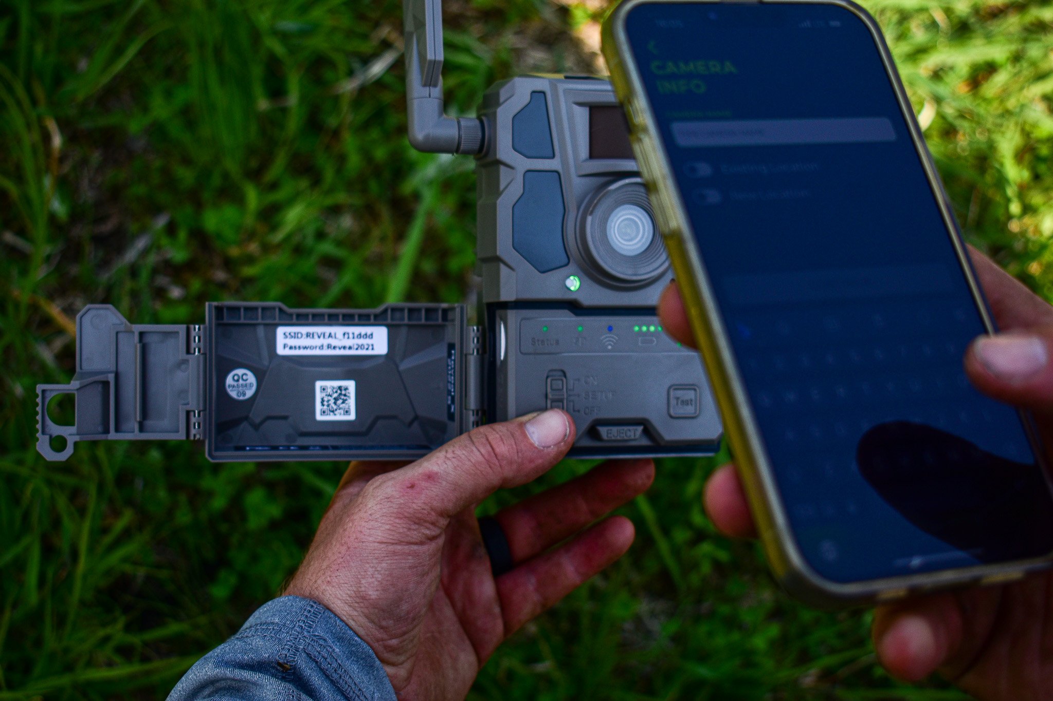 Person setting up a Reveal Cell Camera with a QR code, while also holding a smartphone with a blue screen, outdoors in green grass.