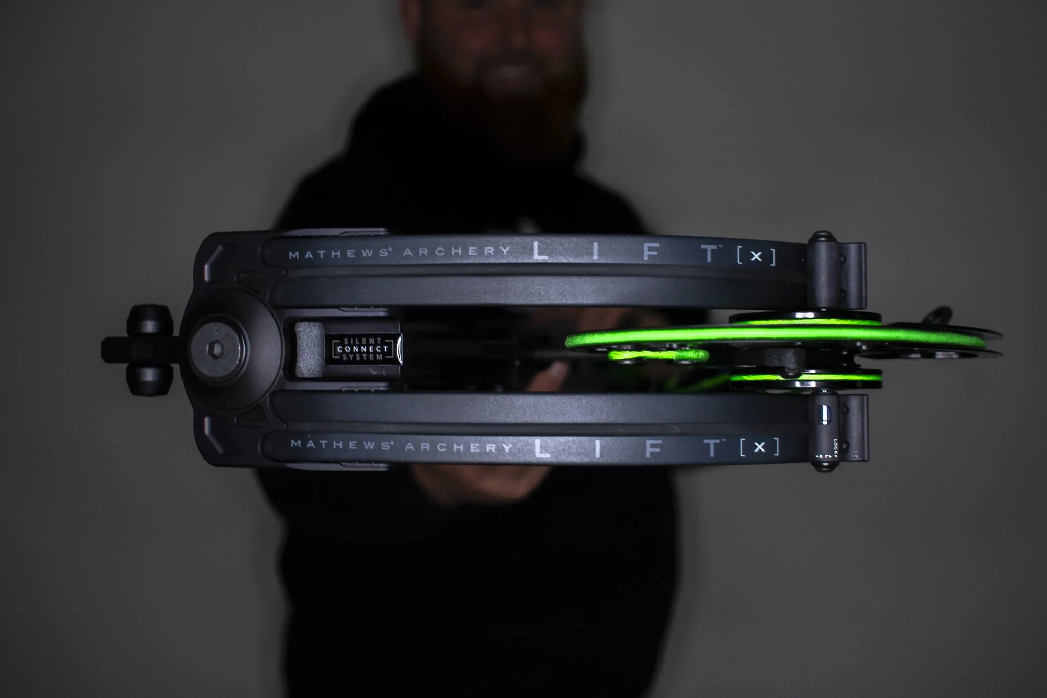 A person holding a Mathews Lift X 33 with green bowstrings, showing the bow's sides and the person's face blurred in the background.