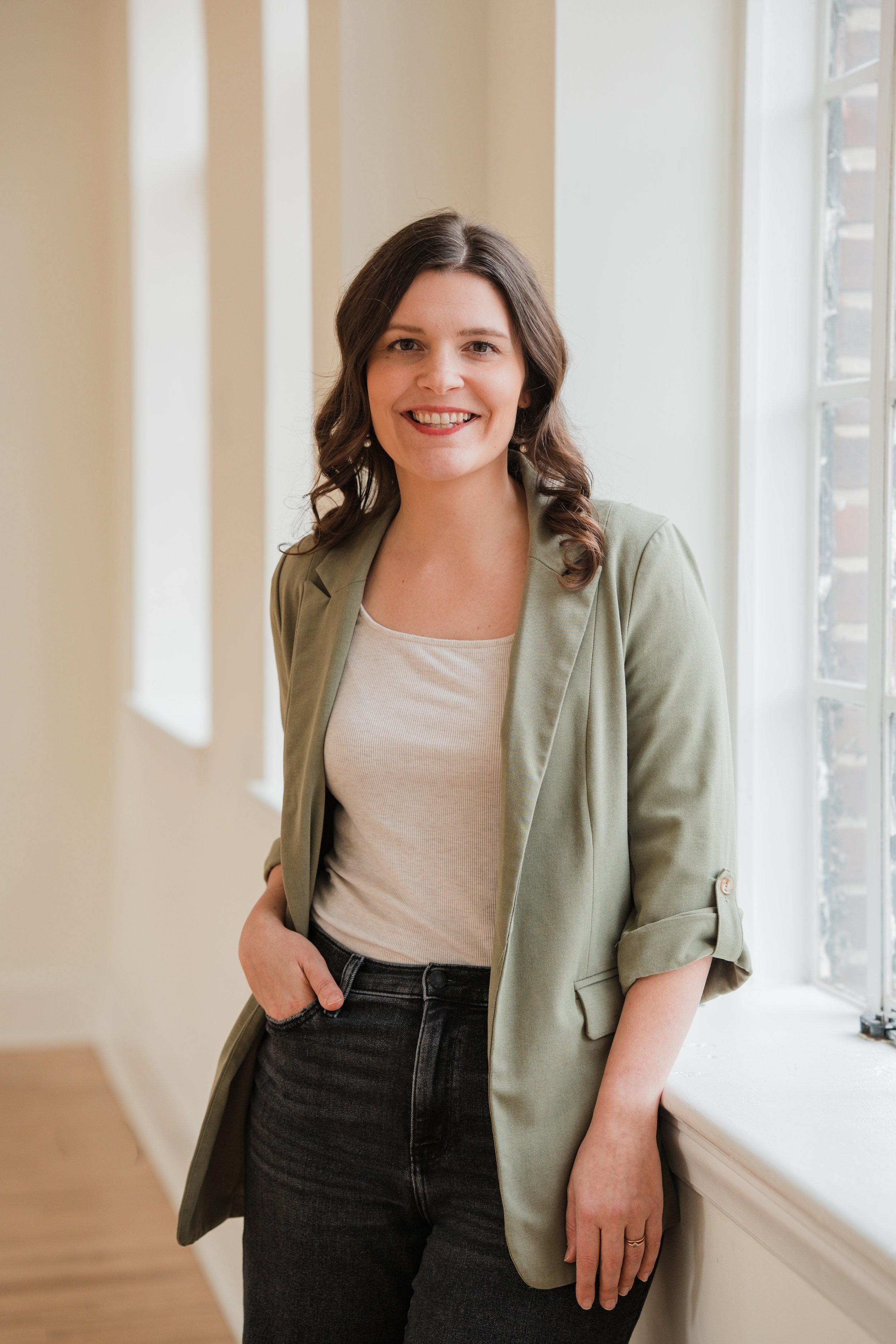 A woman with shoulder-length brown hair wearing a light green blazer, white top, and black jeans, standing indoors next to a window, smiling.
