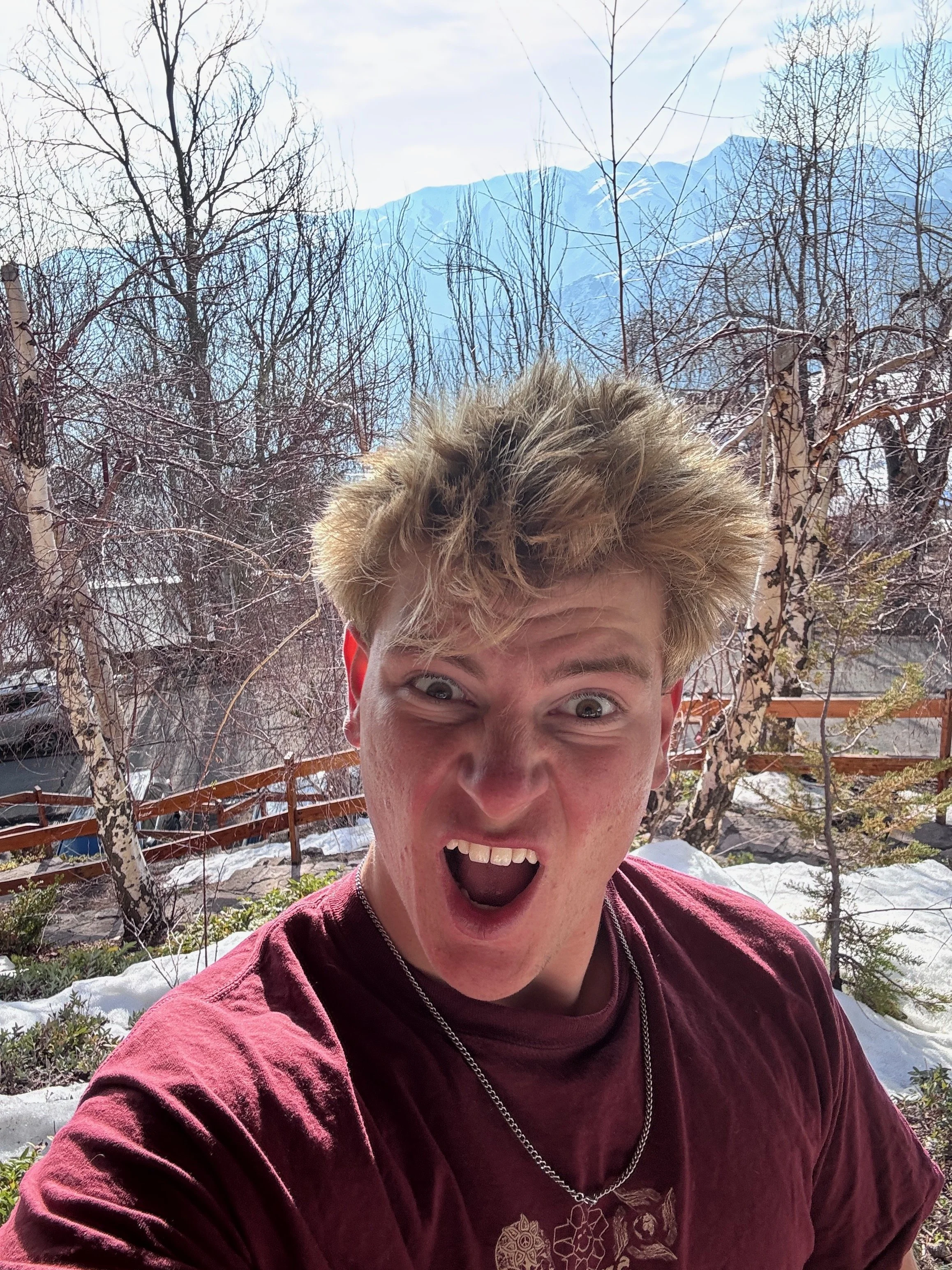A young man with messy blond hair making an excited expression in a snowy outdoor setting with trees and mountains in the background.