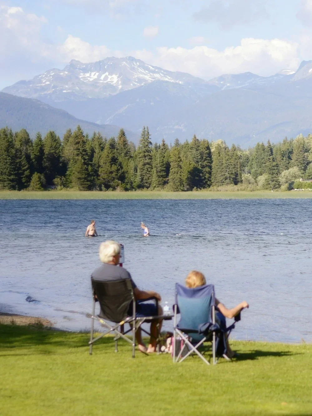 Older man and woman sitting on camping chairs by a lake, with two people swimming in the water and snow-capped mountains and evergreen trees in the background.