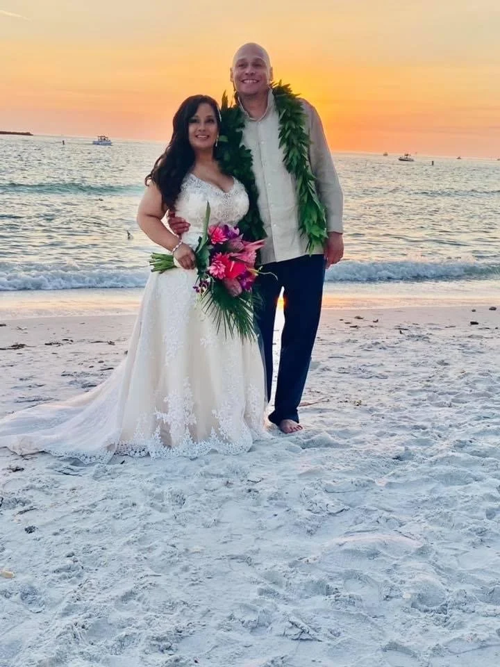 A couple dressed in wedding attire standing on the beach at sunset. The bride is holding a bouquet of pink and purple flowers, and both are wearing green leis. The groom is shirtless with a lei and hugging the bride, smiling at the camera.