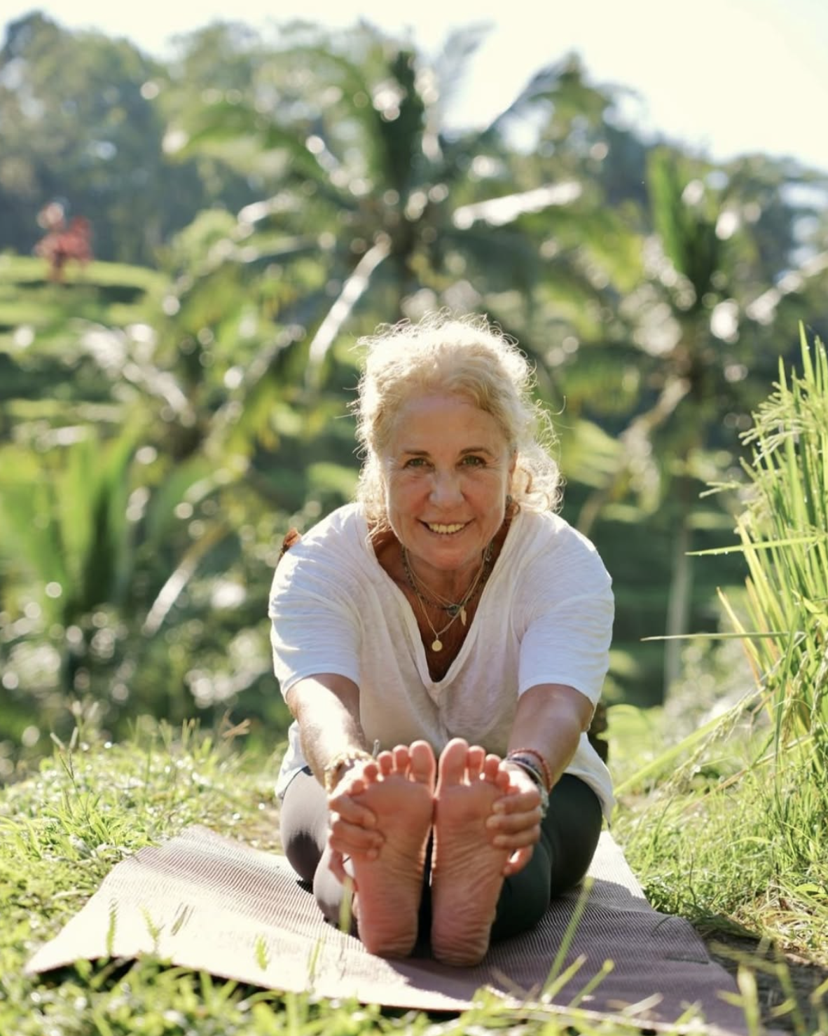 A smiling elderly woman in a white shirt practicing yoga outdoors on a mat in a lush, green garden with trees and plants.