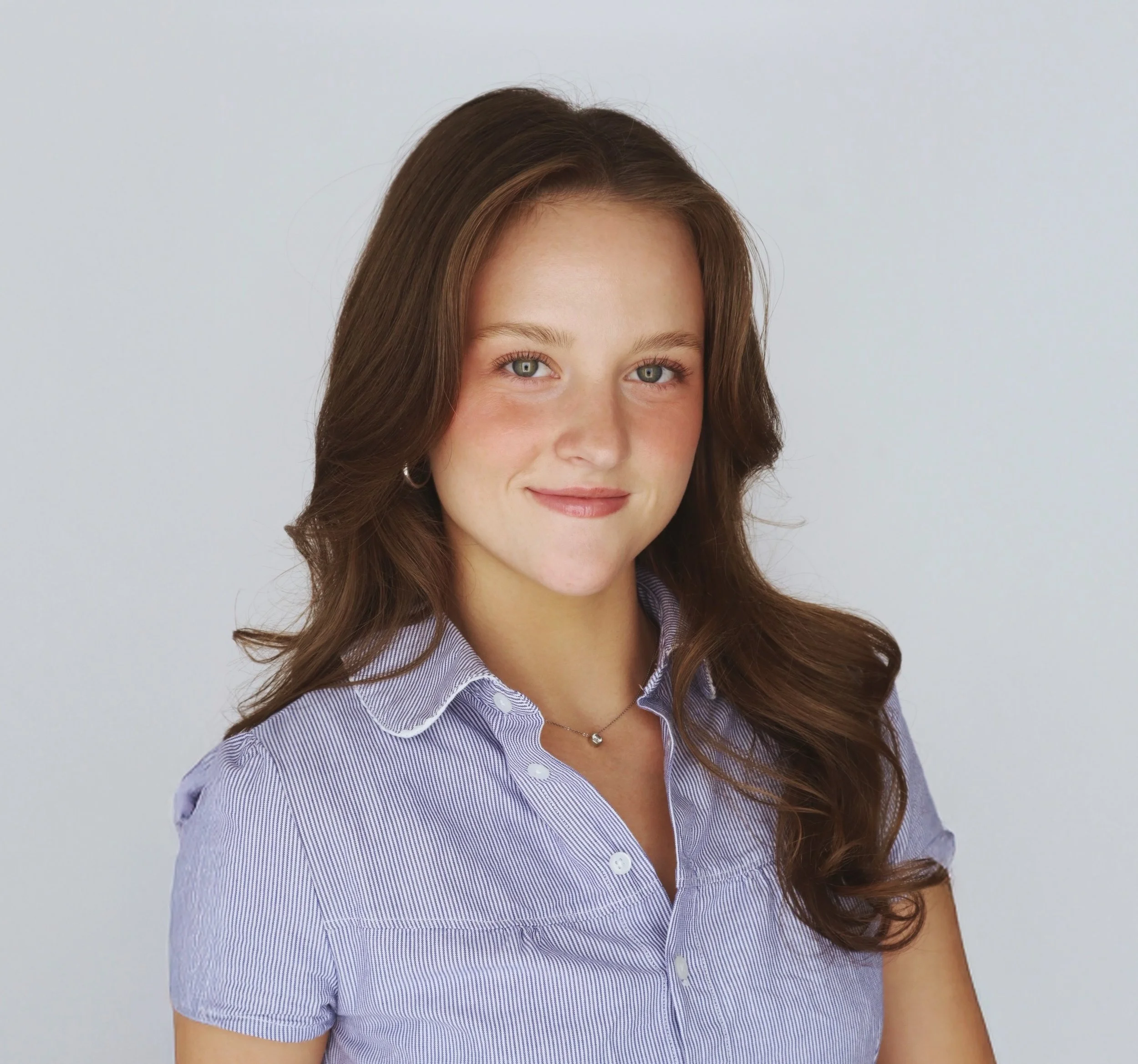 A young woman with long brown hair, wearing a light purple collared shirt and a small necklace, smiling at the camera against a plain light background.