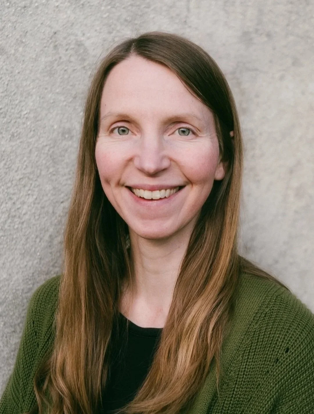 A female therapist with long brown hair, light skin, and green eyes, smiling, wearing a green sweater, standing against a textured light gray wall.