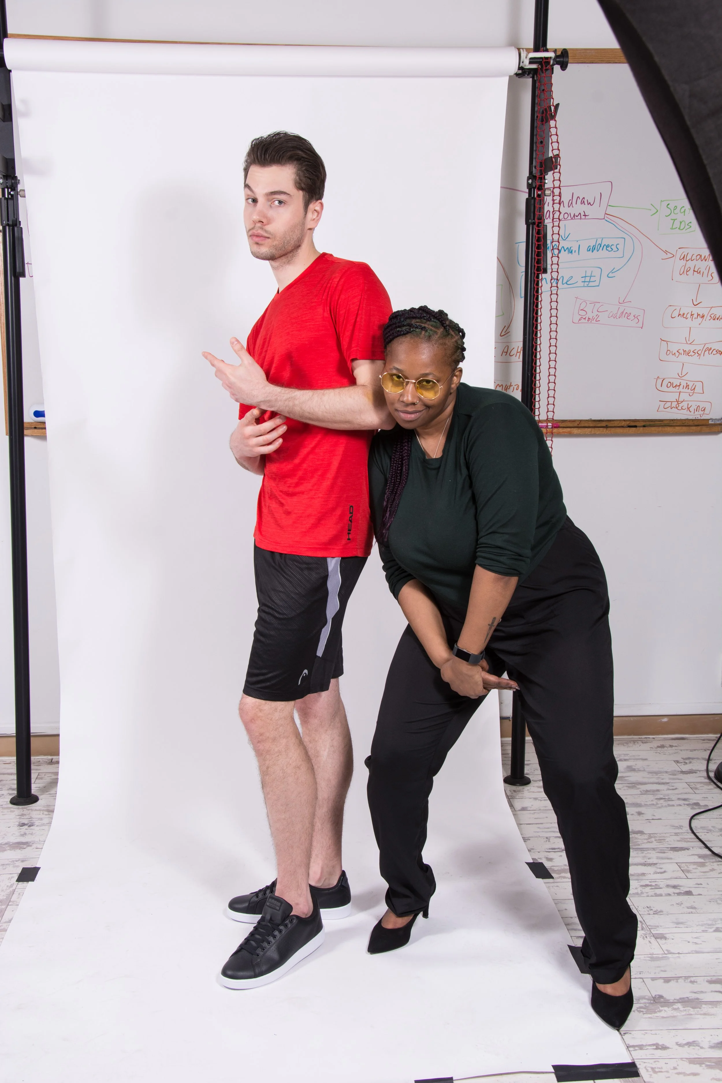 A man and woman posing together in front of a white backdrop, with a whiteboard covered in colorful notes and diagrams in the background. The man is wearing a red t-shirt, black shorts, and black sneakers, and is standing with one hand pointing and the other resting on his stomach. The woman is leaning into the man, wearing a black outfit and yellow-tinted glasses, with her hands clasped in front of her.