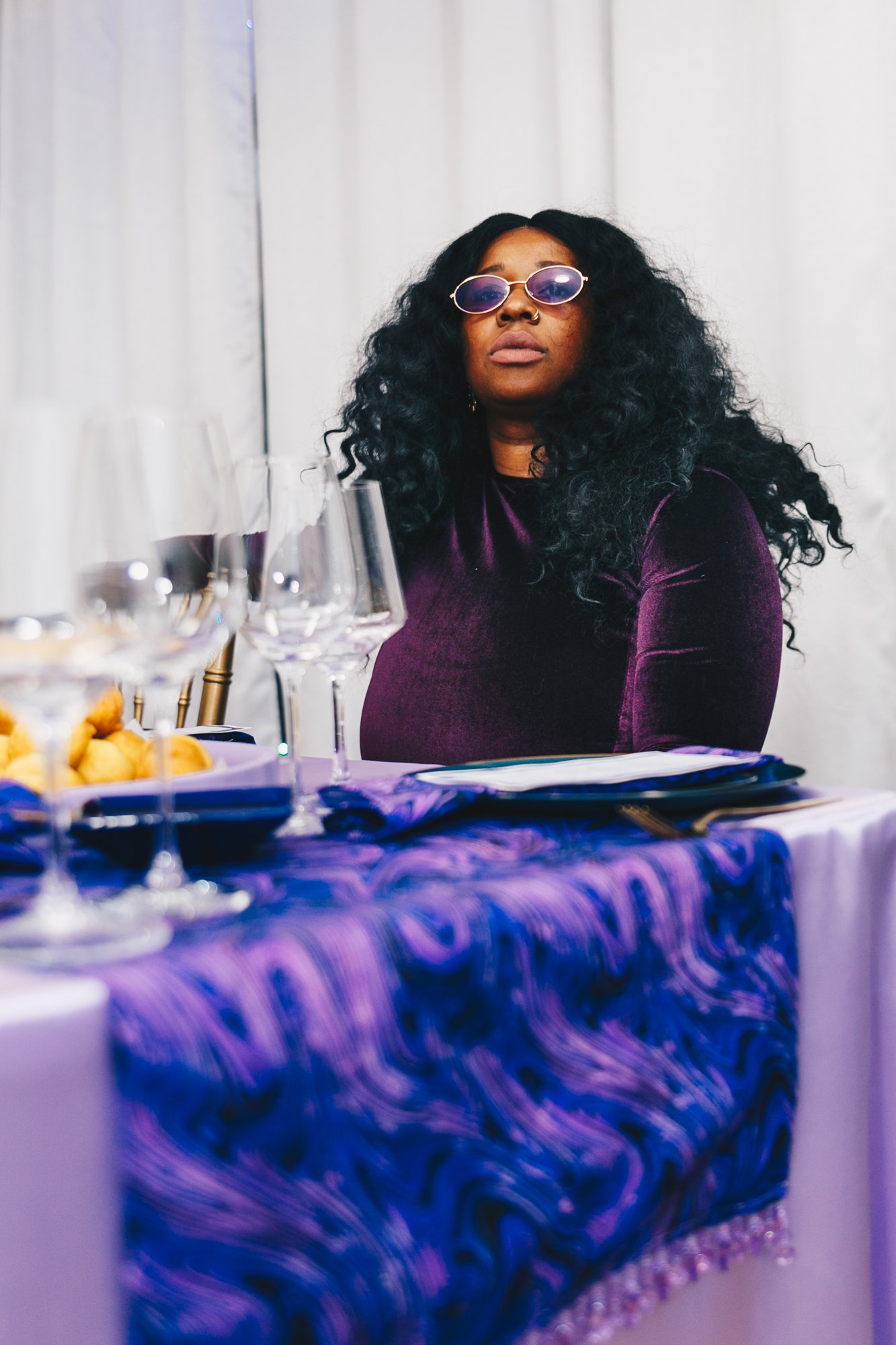 A woman with long, curly black hair wearing purple-tinted sunglasses and a purple velvet top sitting at a table with a purple tablecloth, set with empty wine glasses and a plate of food, against a white curtain background.
