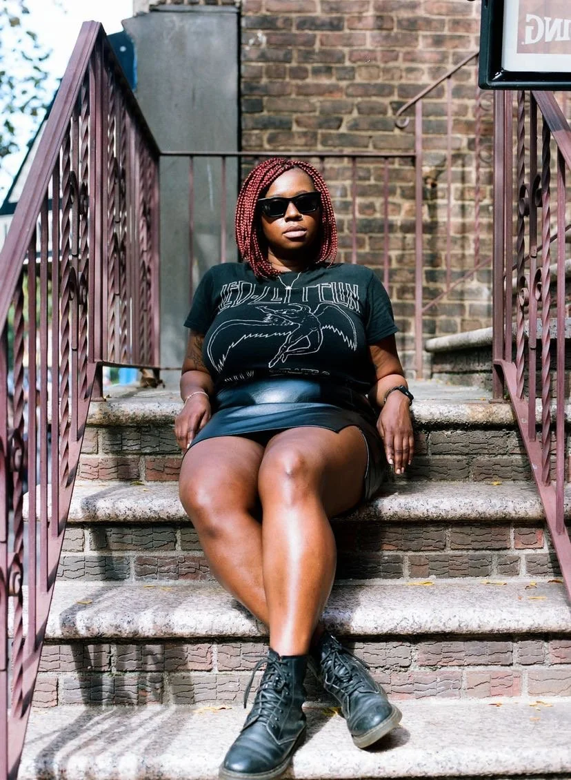 Rae Guy-Thomas photographed on stairs wearing a black leather mini skirt and a Led Zeppelin tee and black cat eye shades