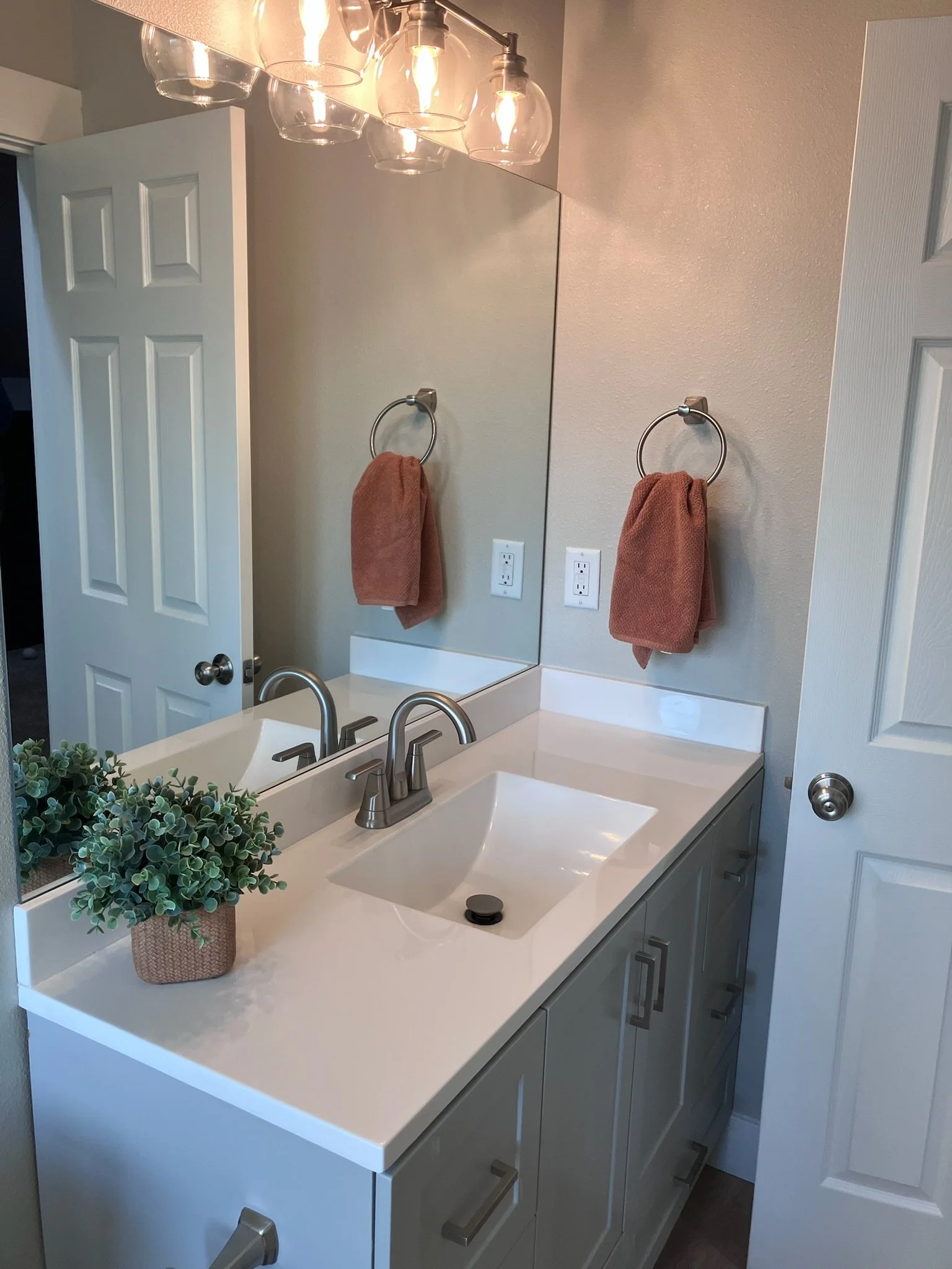 Bathroom vanity with a white sink and dark faucet, a mirror with three lights above, a framed botanical print on the wall, a blue towel on a ring, a small potted plant on the sink, and a bottle of hand soap.