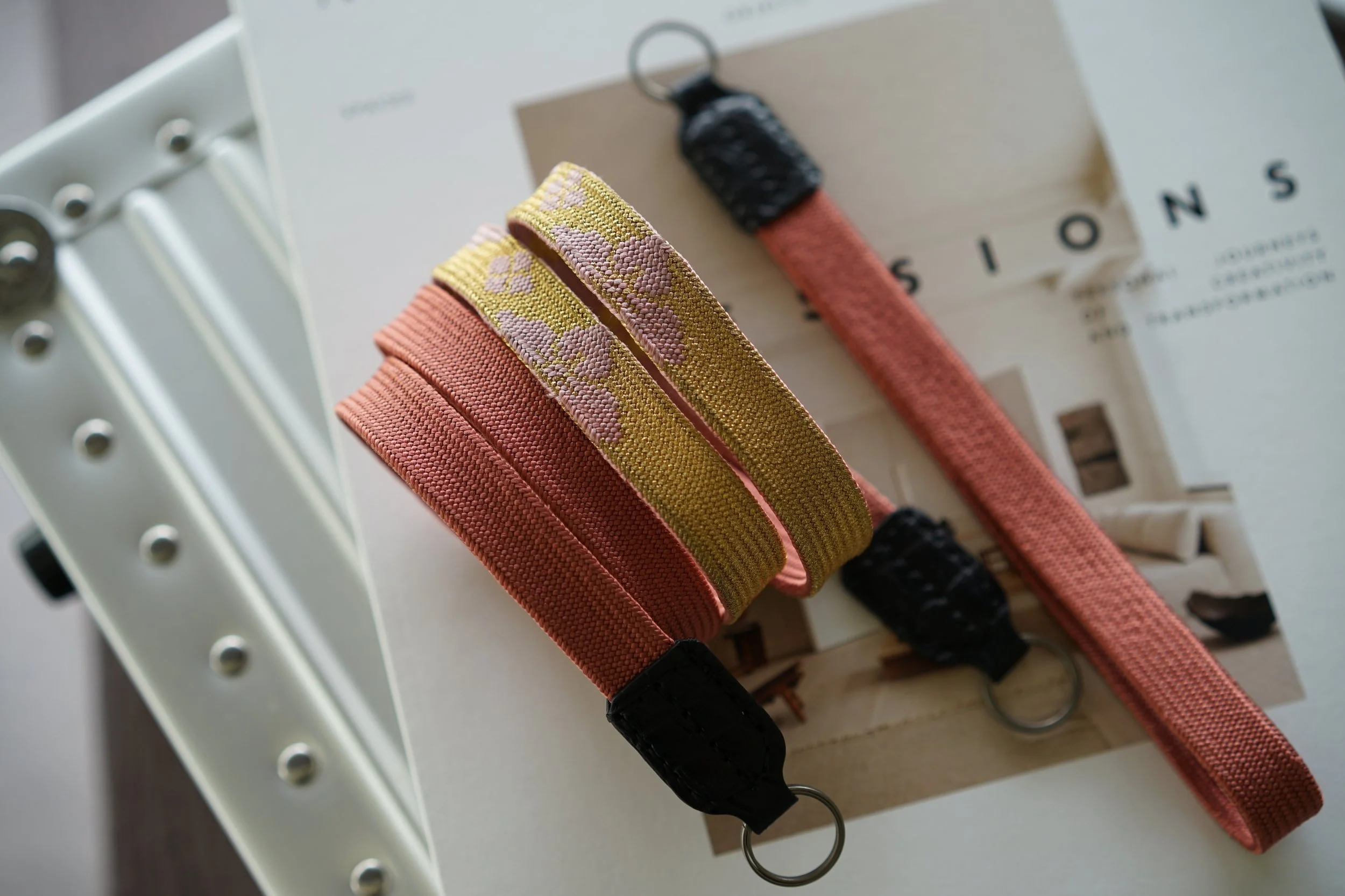 Three fabric keychains with metal rings, placed on top of a magazine or brochure. One keychain is coral, another is yellow with pink floral pattern, and the third is coral with black ends.