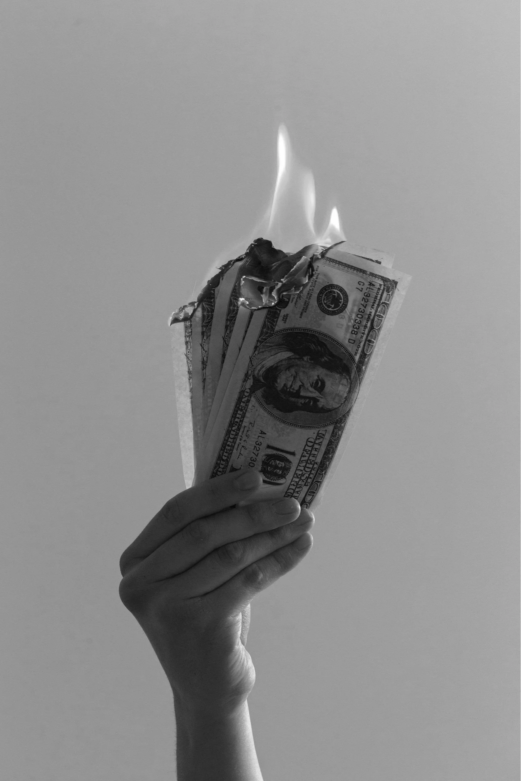 A hand holding a bundle of paper currency, specifically $100 bills, which are on fire.
