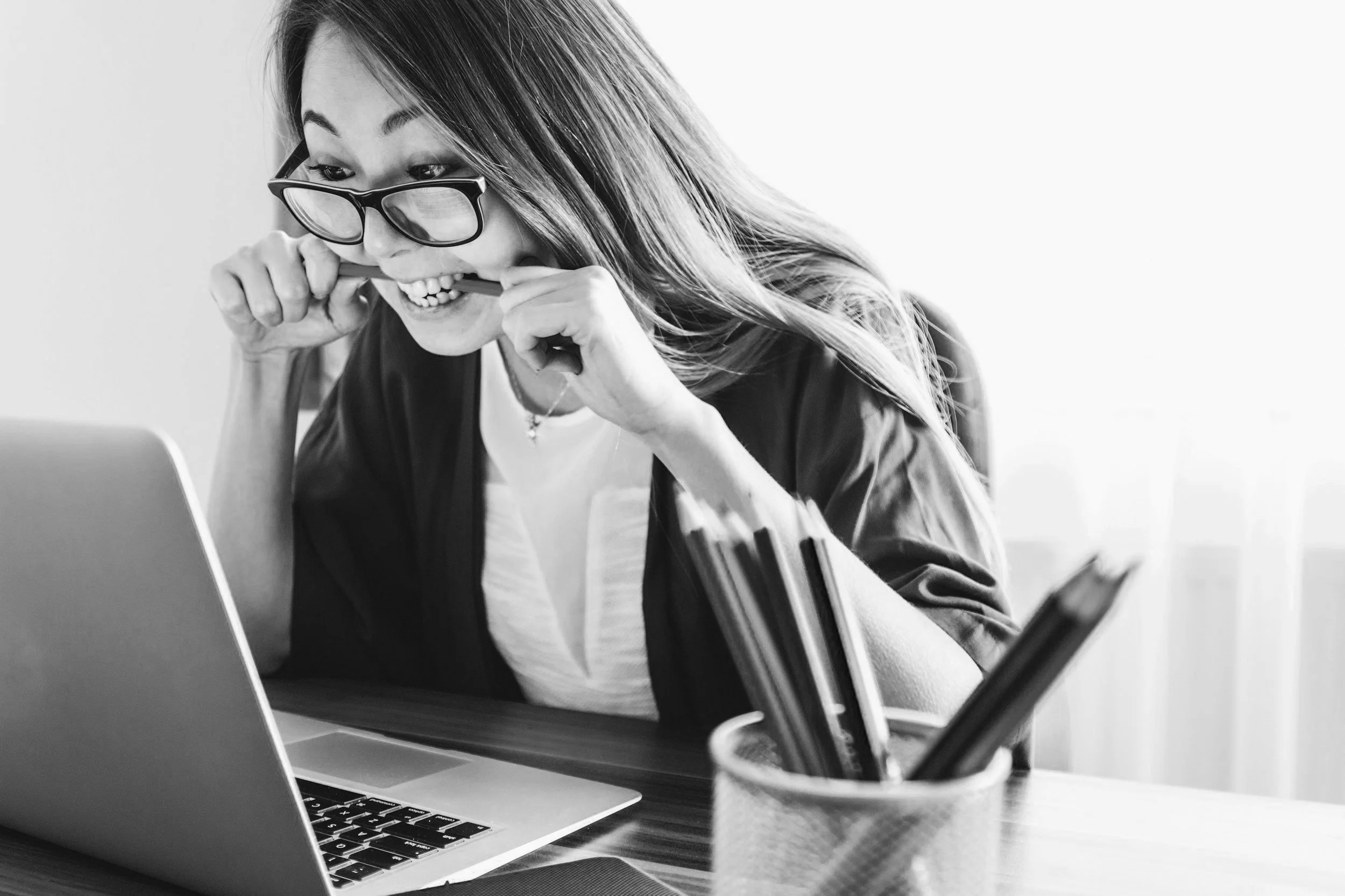 A woman with glasses biting her pencil while looking at her laptop screen, with pens in a holder on the desk.