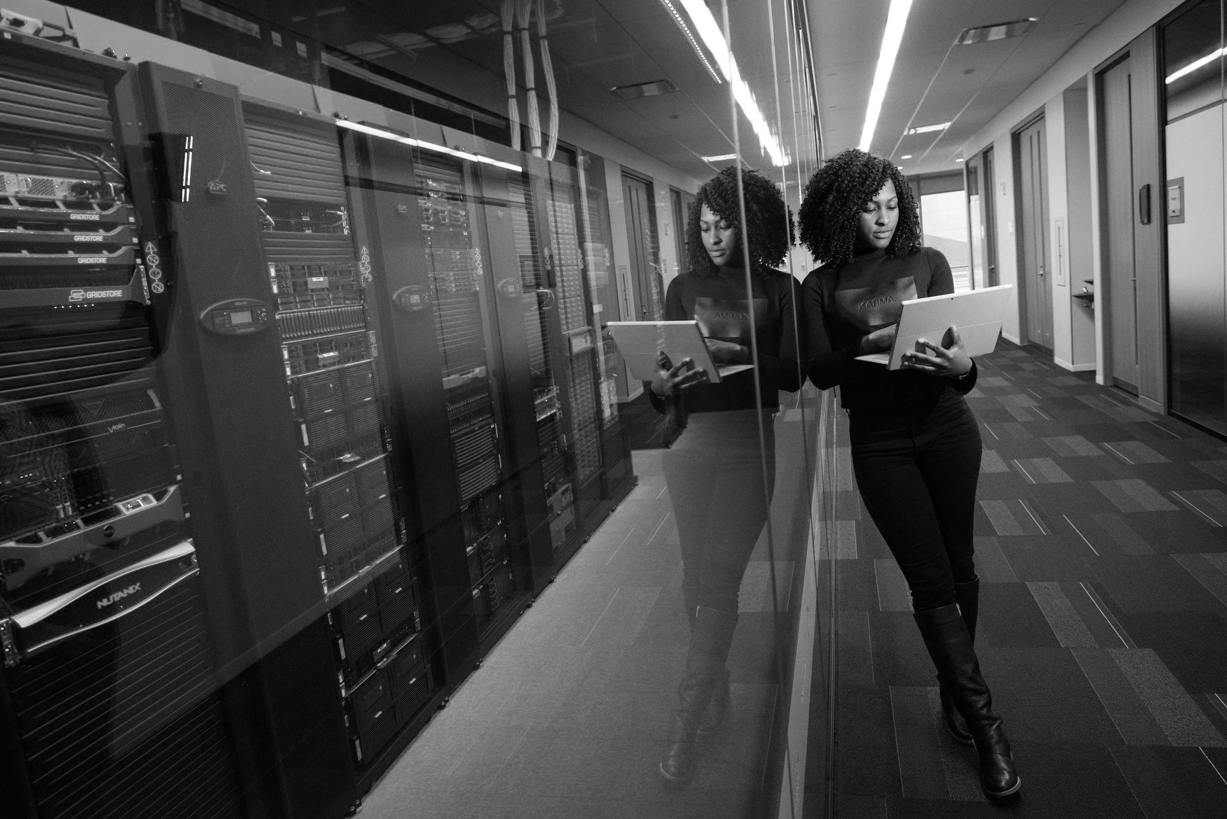 Woman standing next to a glass wall with a reflection, working on a tablet, with server racks in the background.