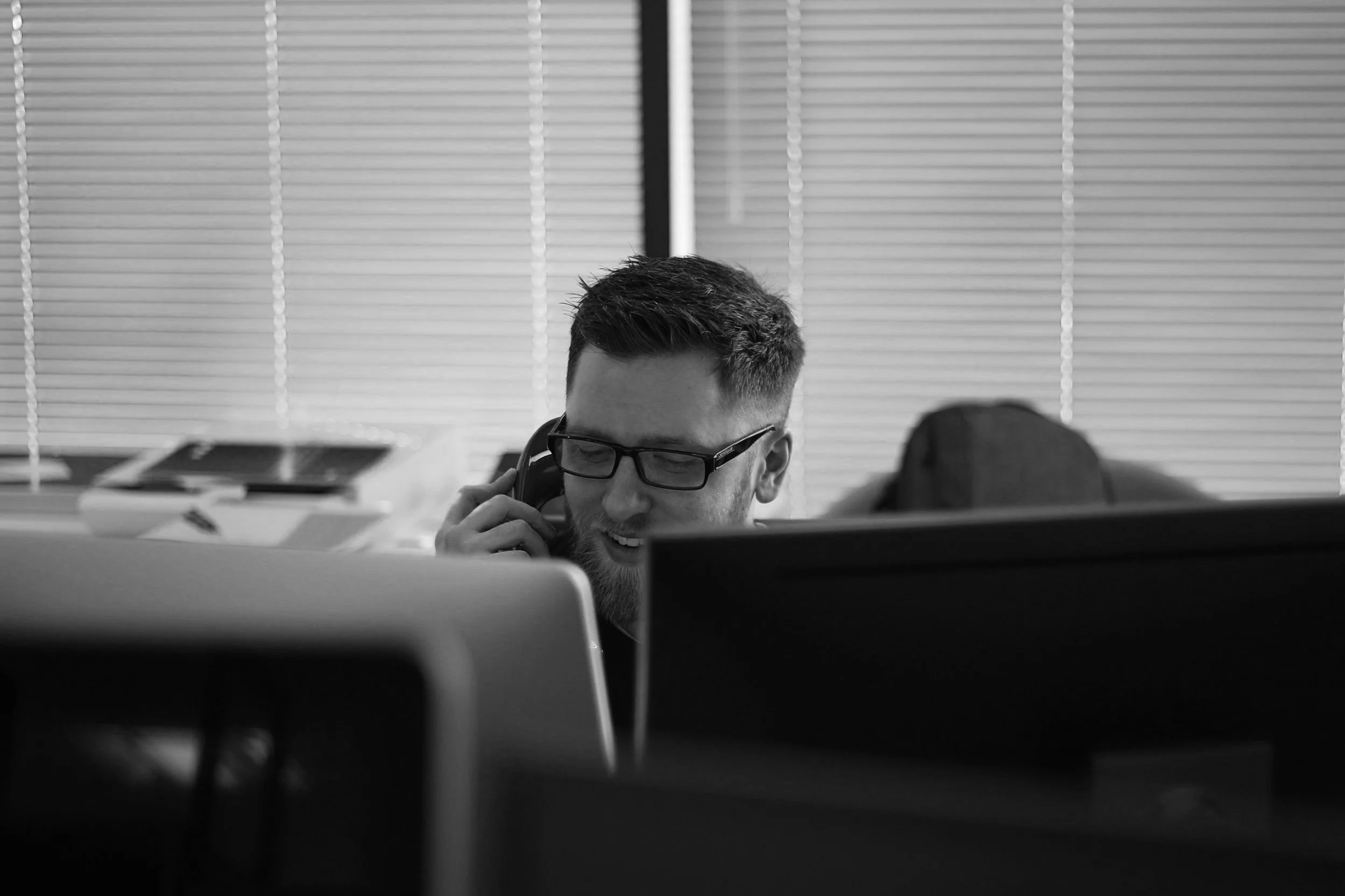 Black and white photo of a man with glasses on a phone, working at a desk with a computer, blinds in the background.
