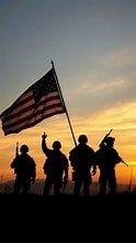 Four soldiers silhouetted against a sunset, one holding an American flag.