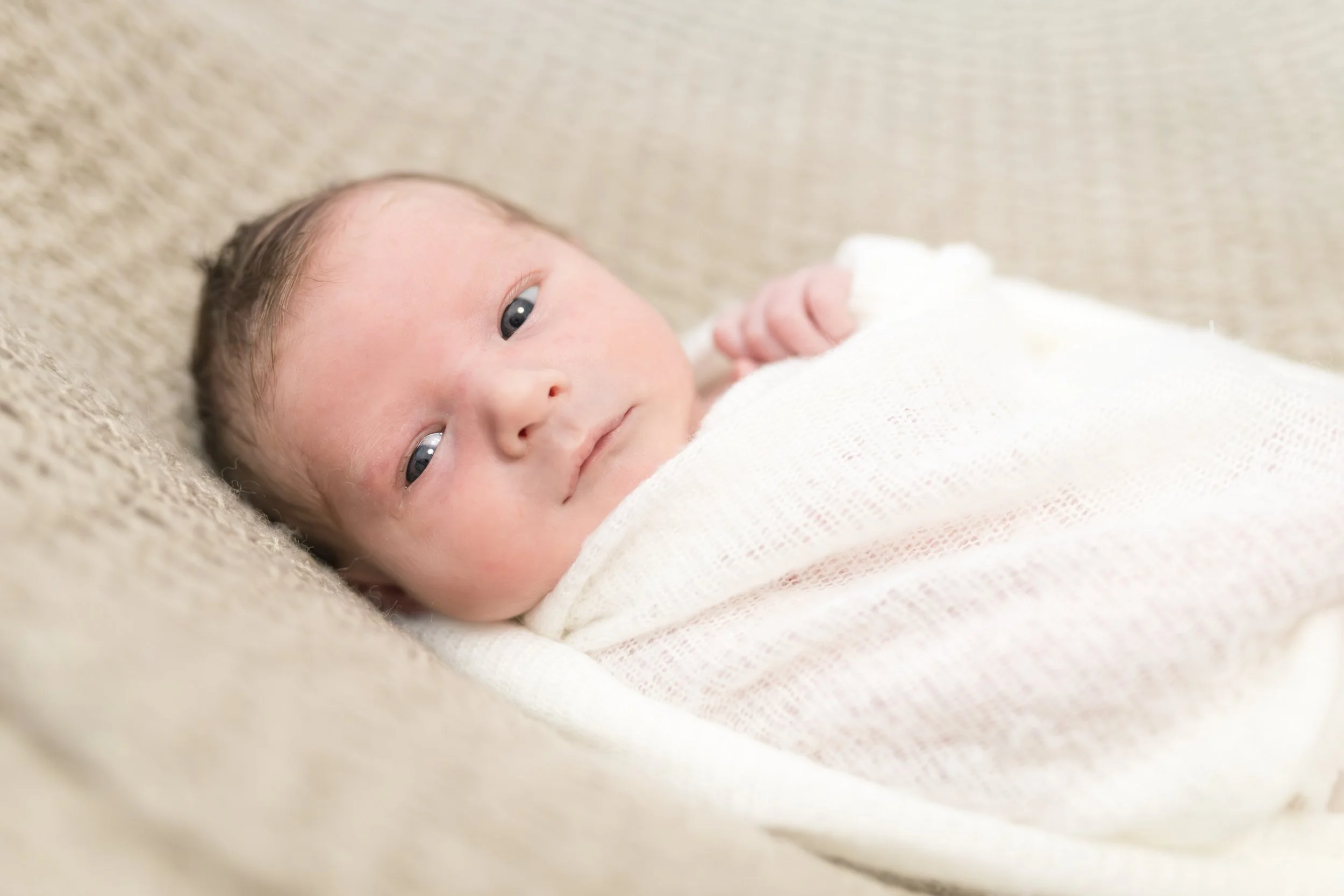 Nahaufnahme eines neugeborenen Babys, das auf einem weichen, beige-farbenen Bezug liegt, mit blauen Augen und einem weißen Strickpullover, das Baby schaut direkt in die Kamera.