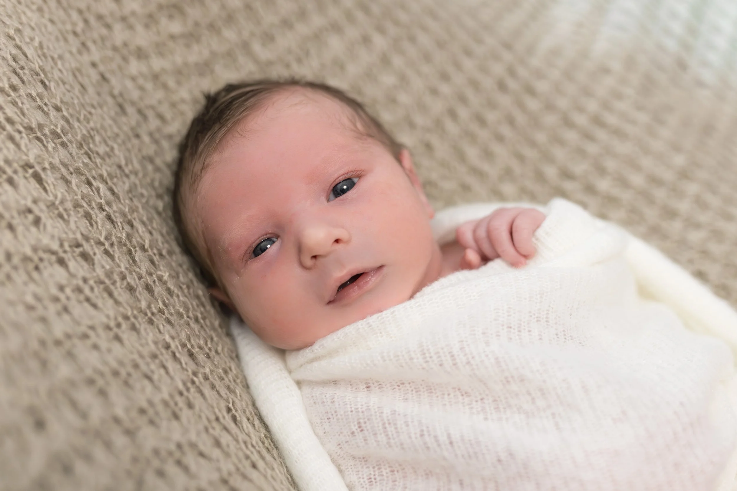 Nahaufnahme eines neugeborenen Babys, das auf einer beige Textur liegt und hell gekleidet ist.