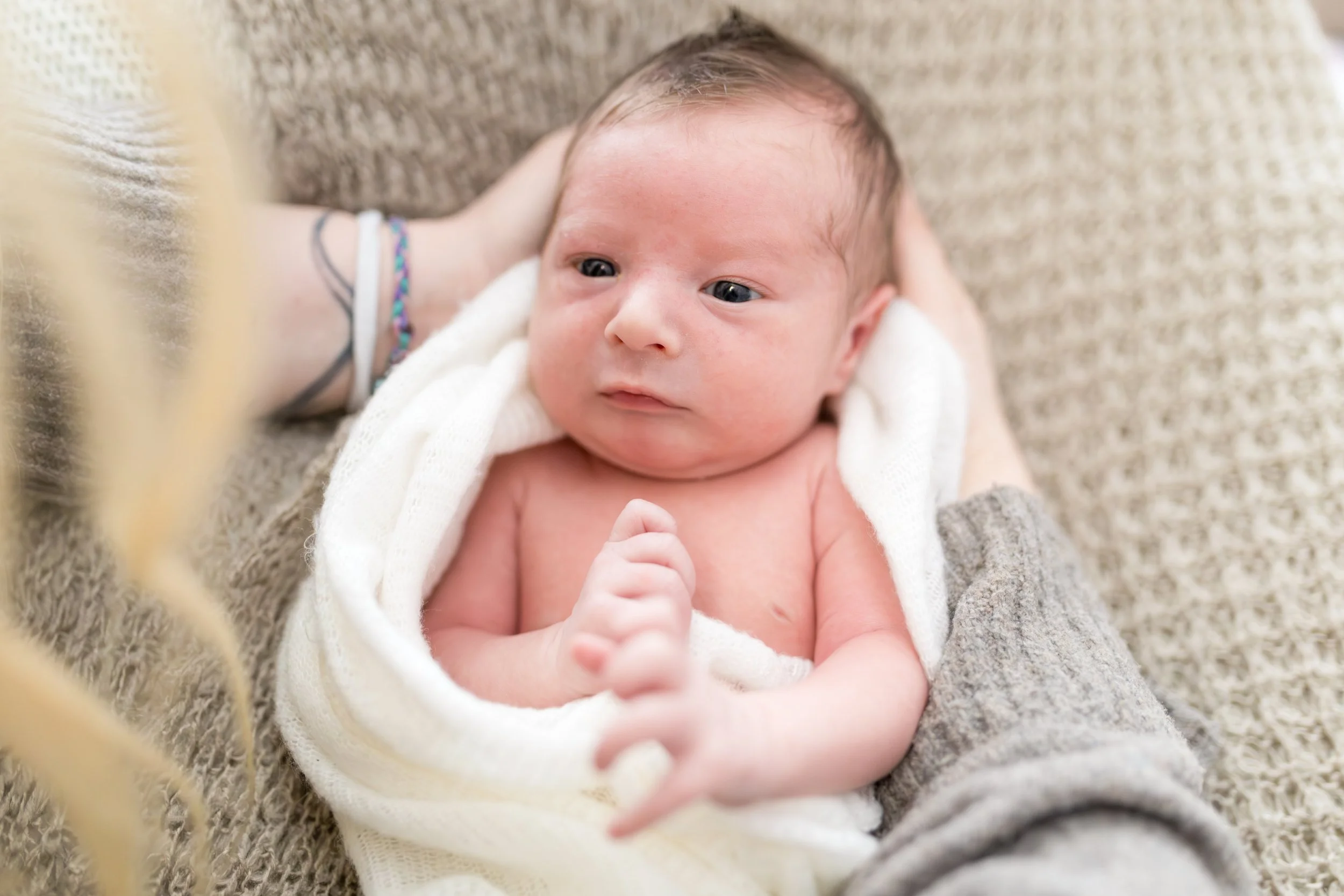 Nahaufnahme eines neugeborenen Babys, das auf einer weichen, grauen Decke liegt, mit Blick nach oben, umgeben von zarten, weißen Tüchern.