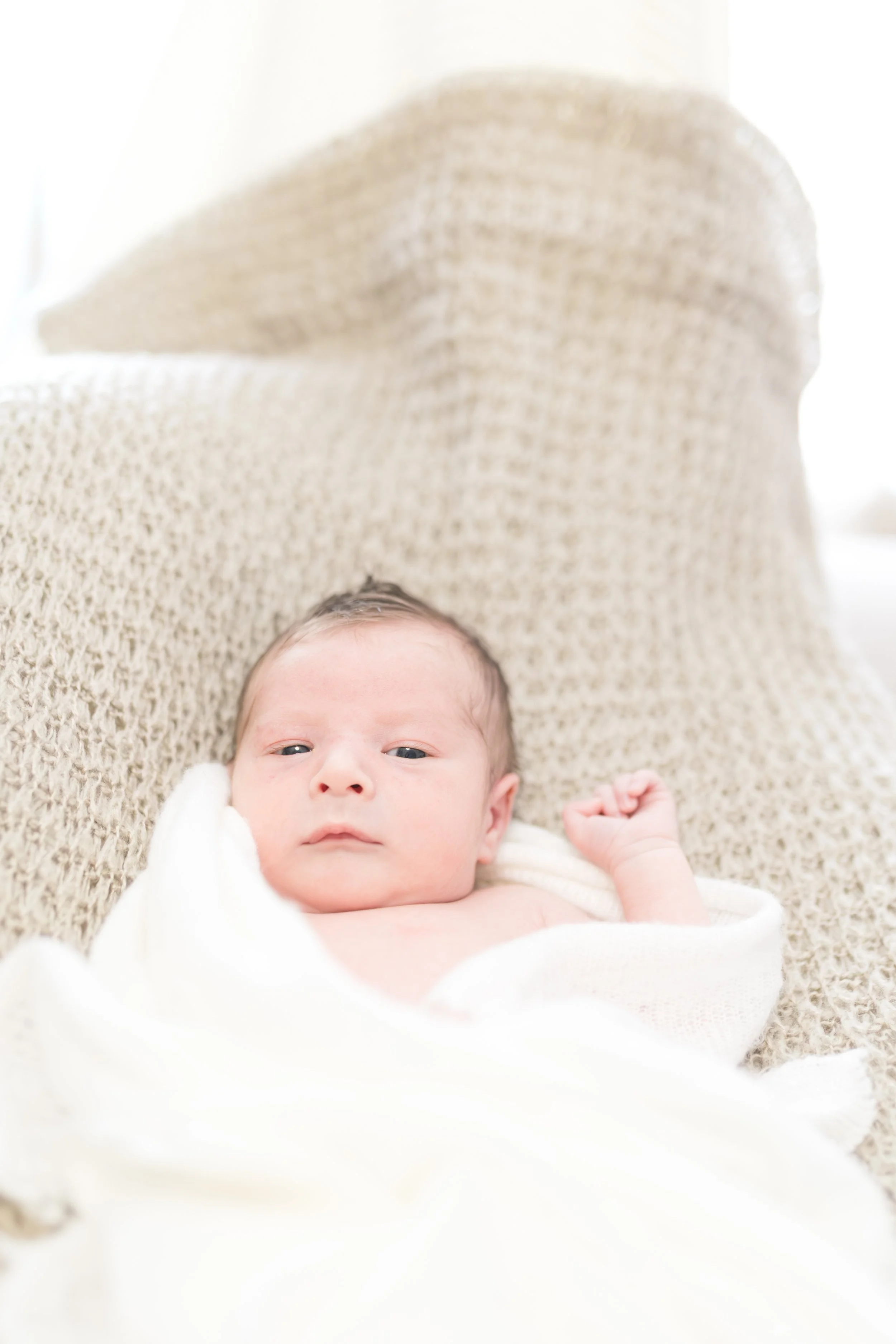 Ein neugeborenes Baby liegt auf einer weißen Decke, eingewickelt in ein cremefarbenes Tuch, mit einem hellen, wollweißen Kissen im Hintergrund.