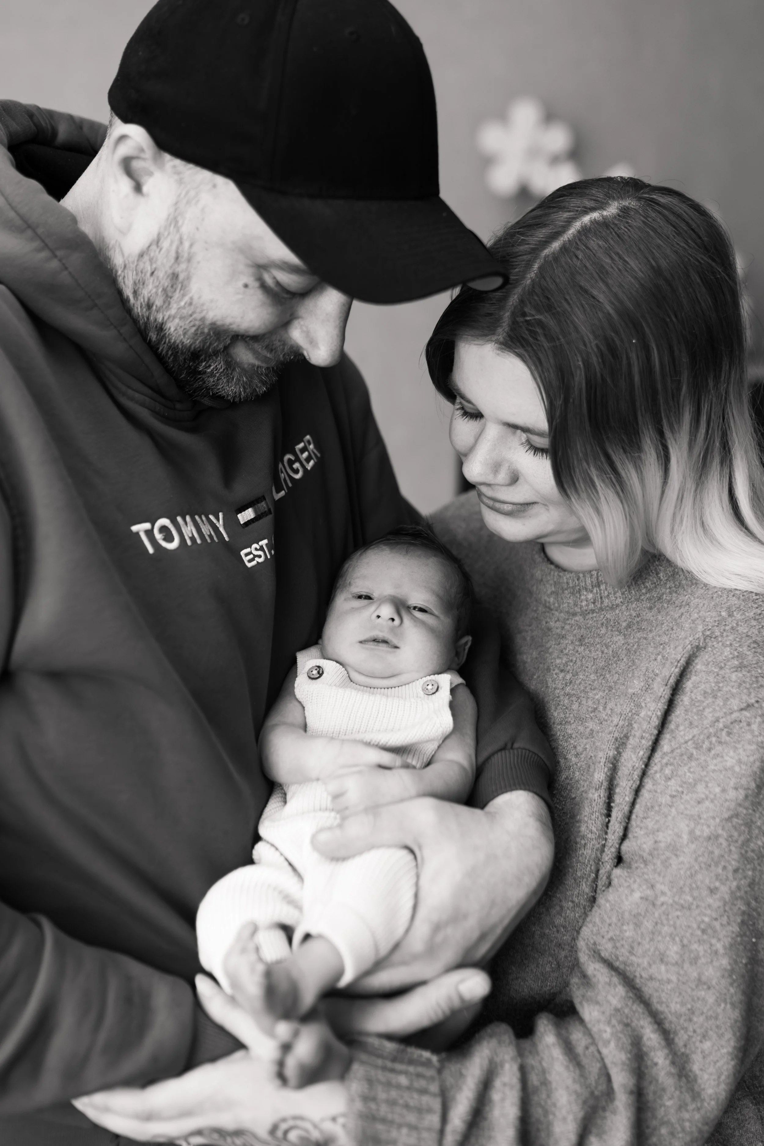 Ein Mann und eine Frau halten gemeinsam ein Baby in den Armen. Das Gesicht des Babys ist sichtbar, während die Erwachsenen sich liebevoll zu ihm beugen. Das Bild ist in Schwarz-Weiß aufgenommen.