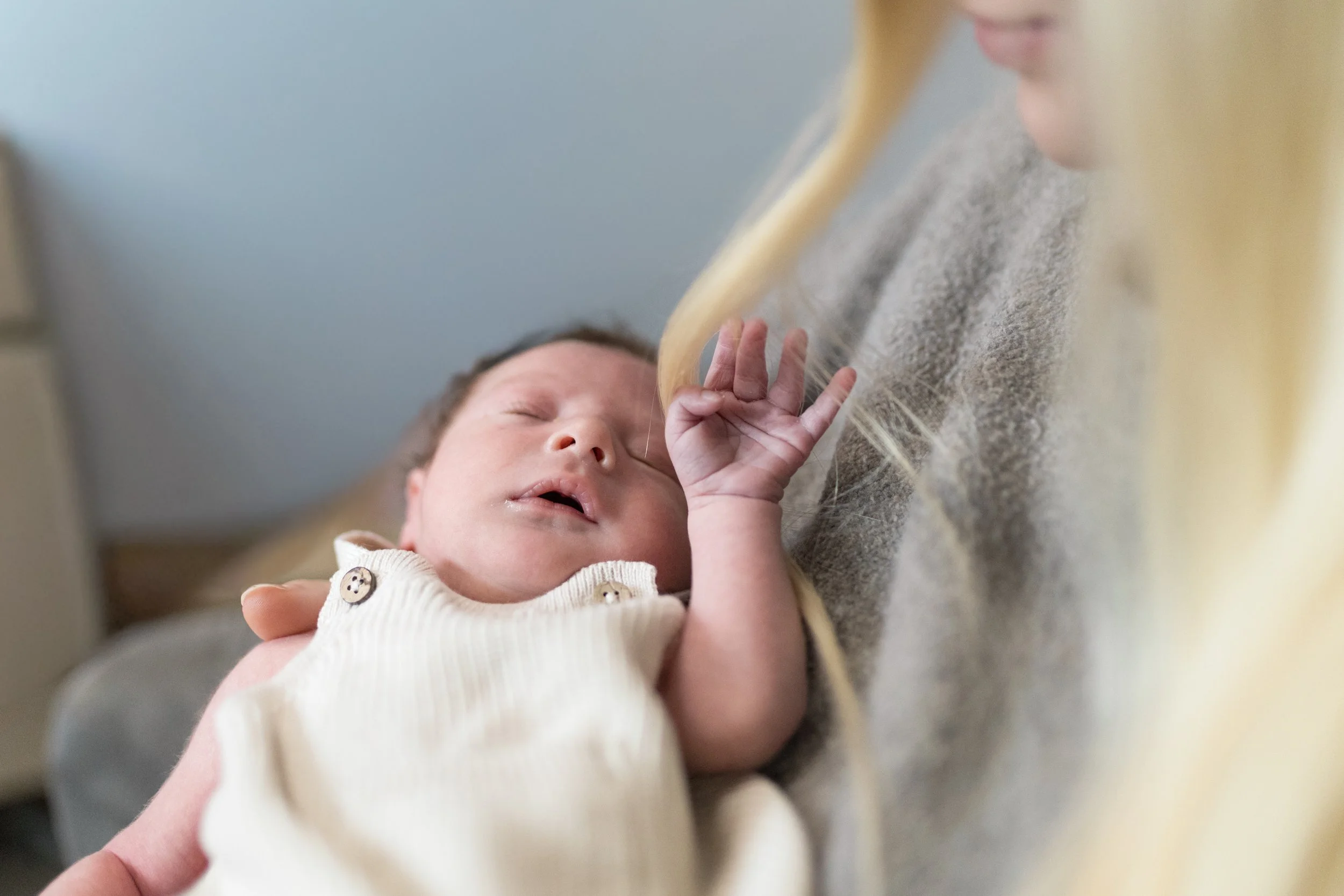 Ein Baby liegt in den Armen einer Frau, die es sanft hält, während es schläft. Das Baby trägt ein cremefarbenes Kleid mit Knöpfen, und die Szene ist warm und liebevoll.