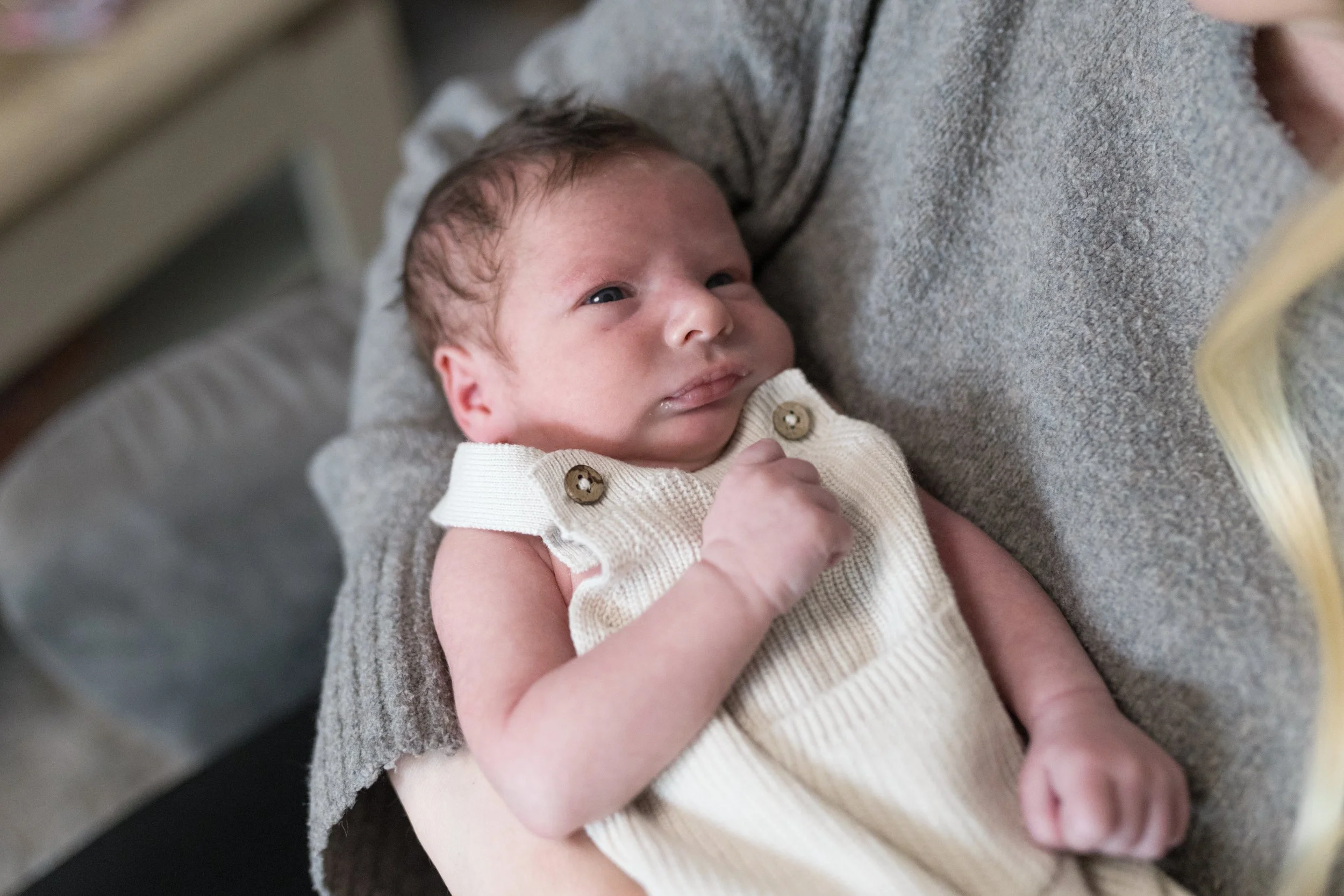 Kleiner Baby mit hellbraunen Haaren, liegend auf einer grauen Decke, trägt eine beigefarbene Strickweste und schaut nach oben.