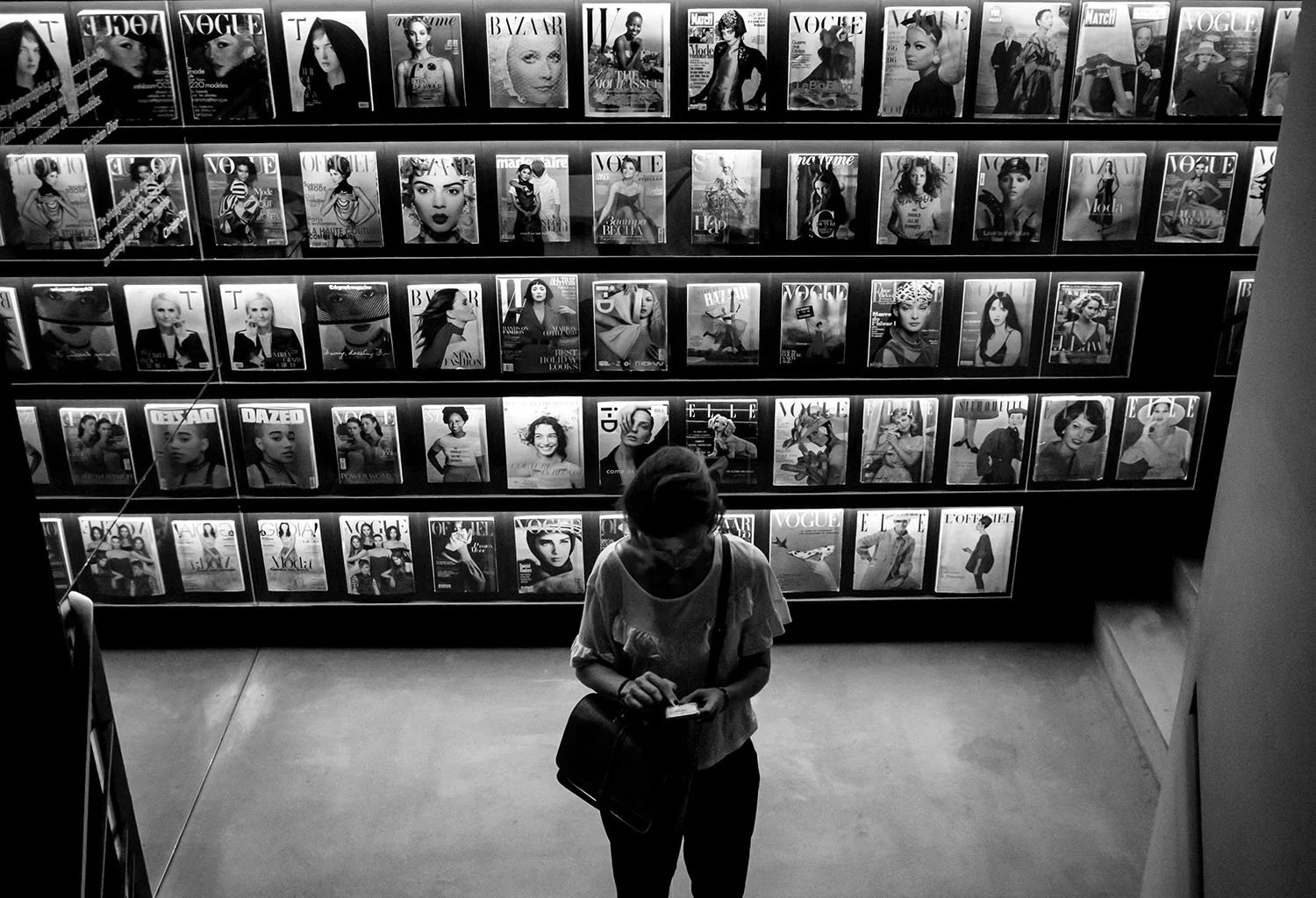 In monochrome silence, a visitor stands beneath a wall of fashion imagery that shaped the visual language of couture.