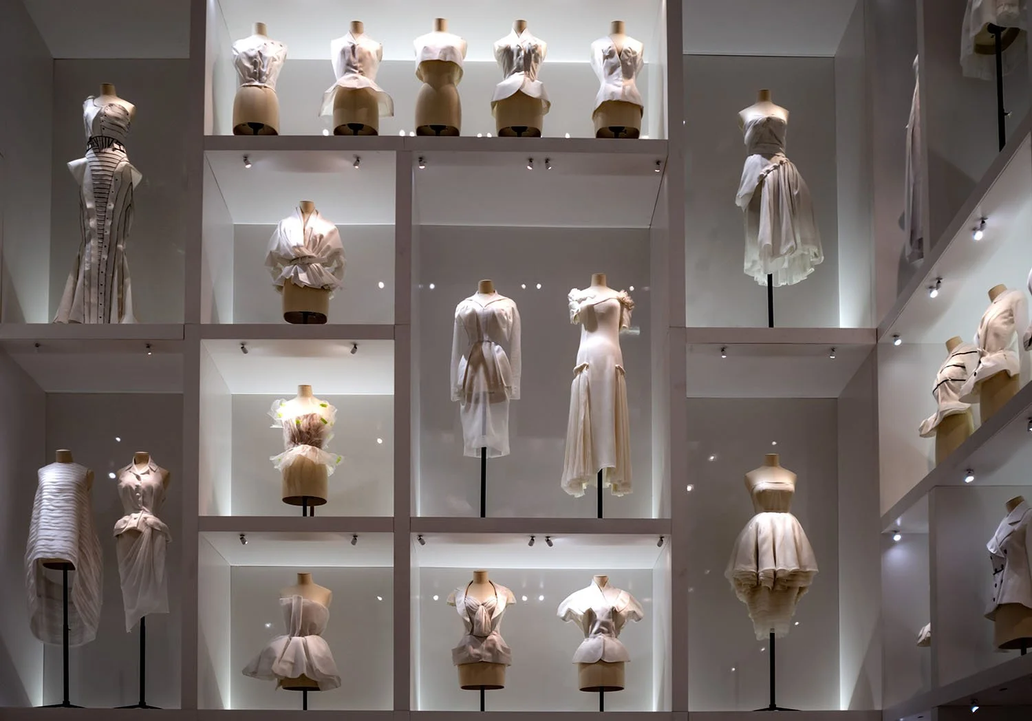 A wall of illuminated compartments presents a study of Dior silhouettes, revealing variations in form, structure, and couture design.