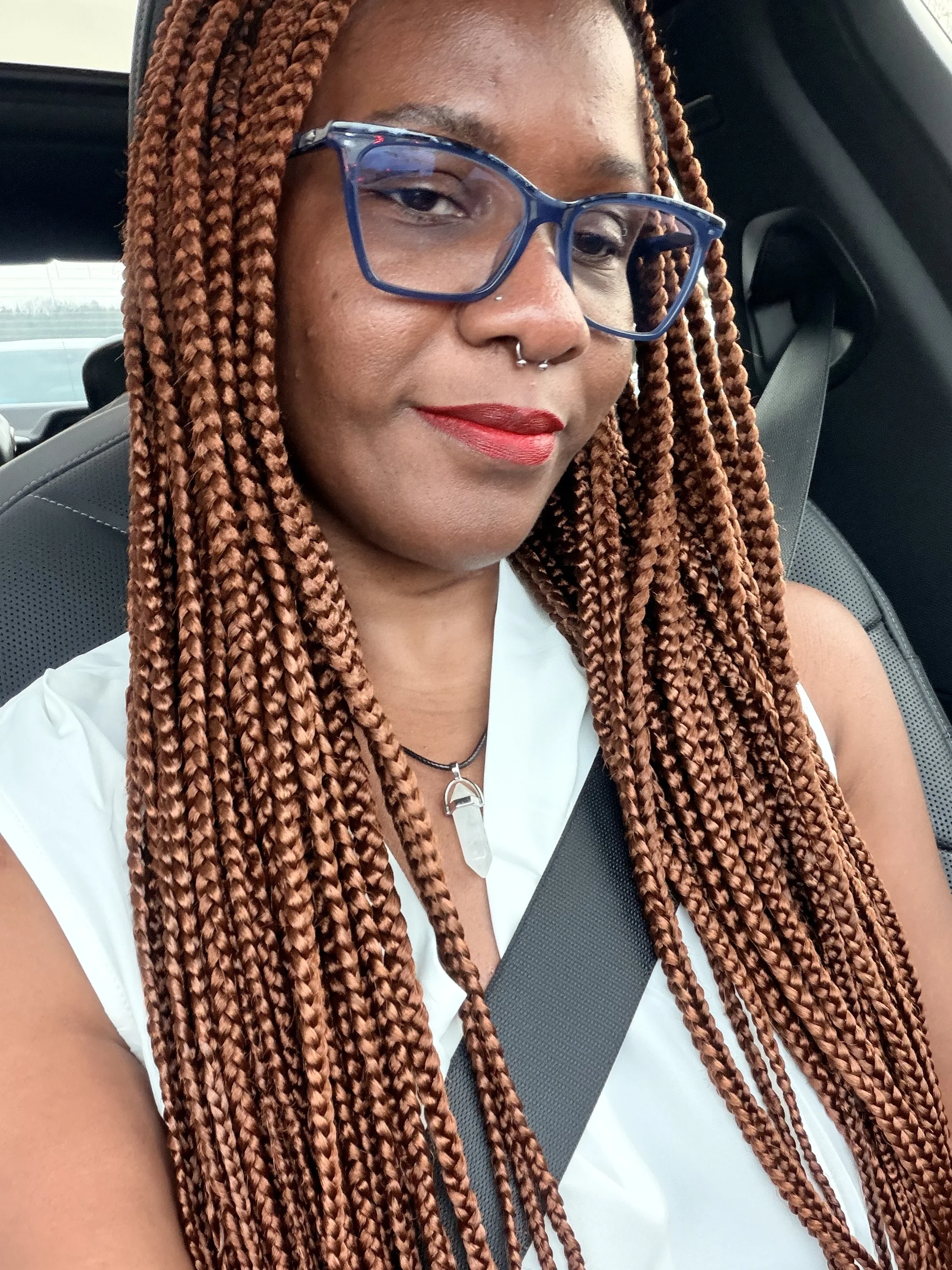 A person with long, braided hair wearing blue glasses, a white top, and a septum piercing, sitting in a car with a seatbelt on.