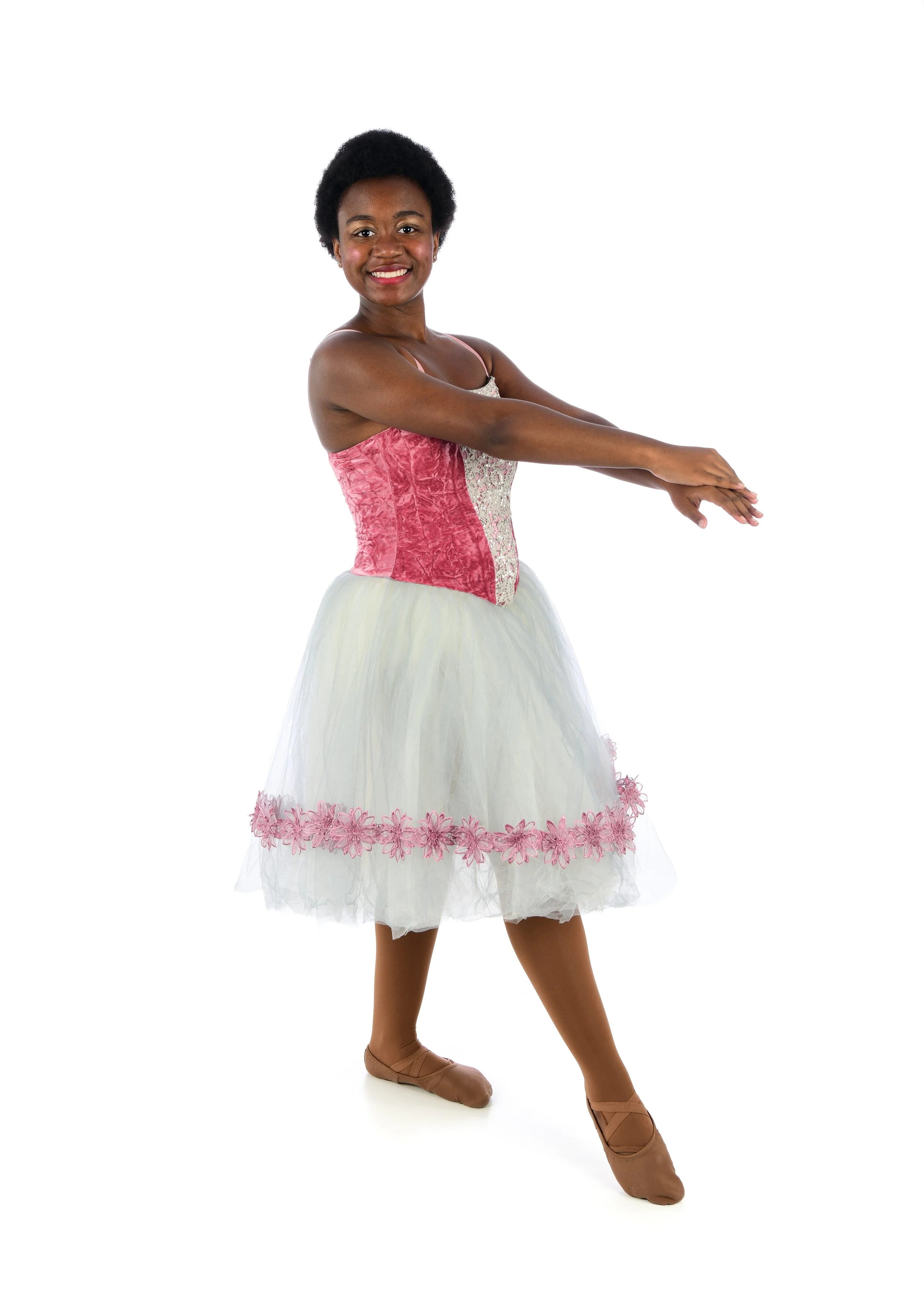 A young woman with dark skin and short curly black hair is posing in a ballet pose. She is wearing a pink and white ballet tutu with floral lace accents, pink tights, and tan ballet slippers. She is smiling and extending her arms forward.