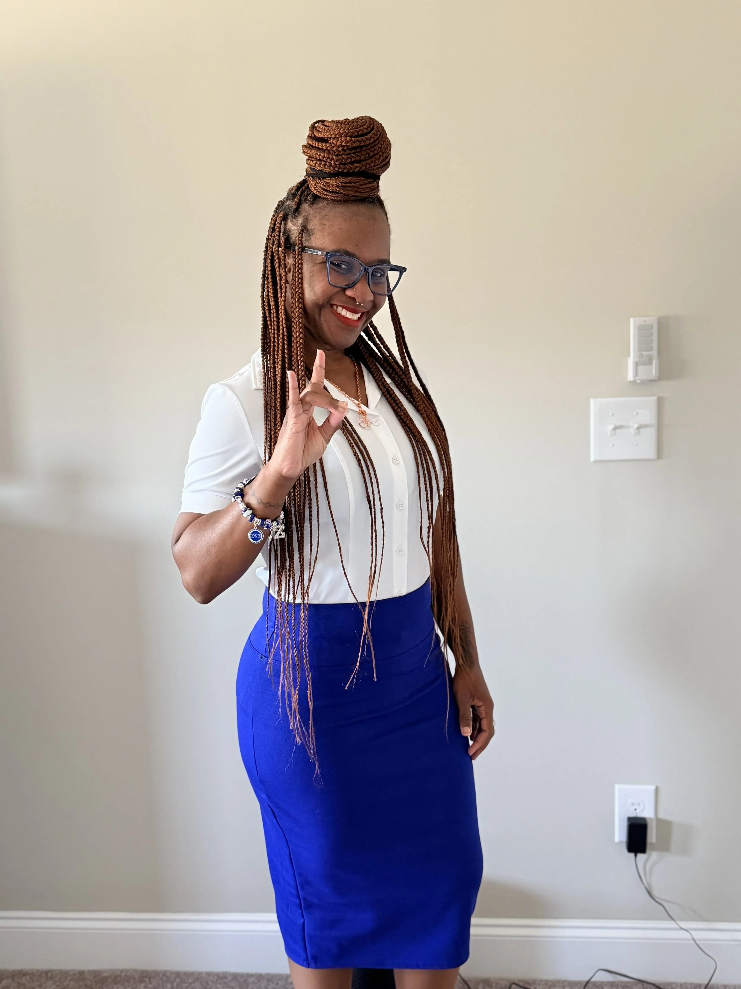 A smiling woman with braided hair styled in a high bun, wearing glasses, a white blouse, and a blue skirt, making an okay gesture with her hand against a plain wall background.