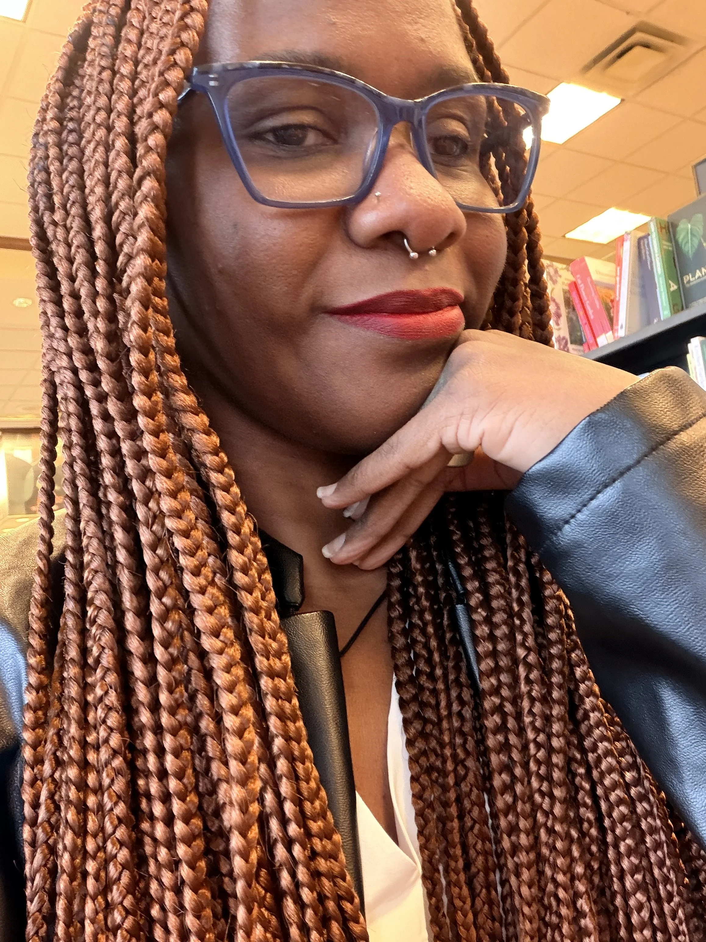 Close-up of a woman with braided hair, wearing glasses and a leather jacket, sitting in a library or bookstore.
