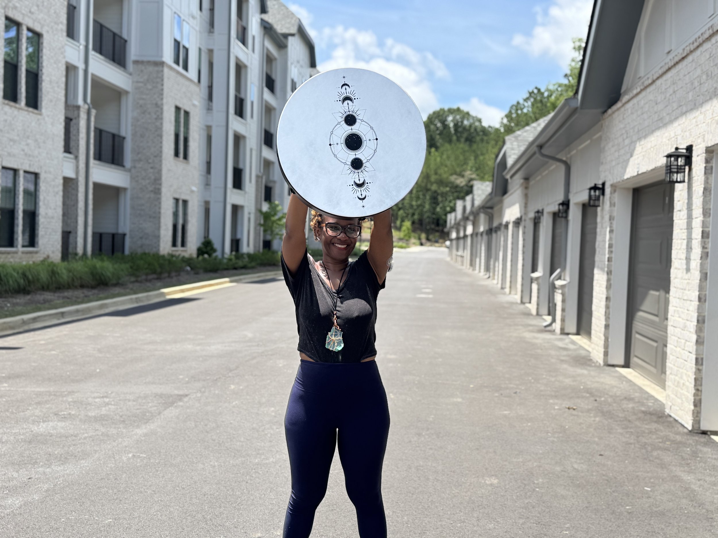 A smiling woman holding a circular object with moon and celestial designs above her head in a residential alleyway.