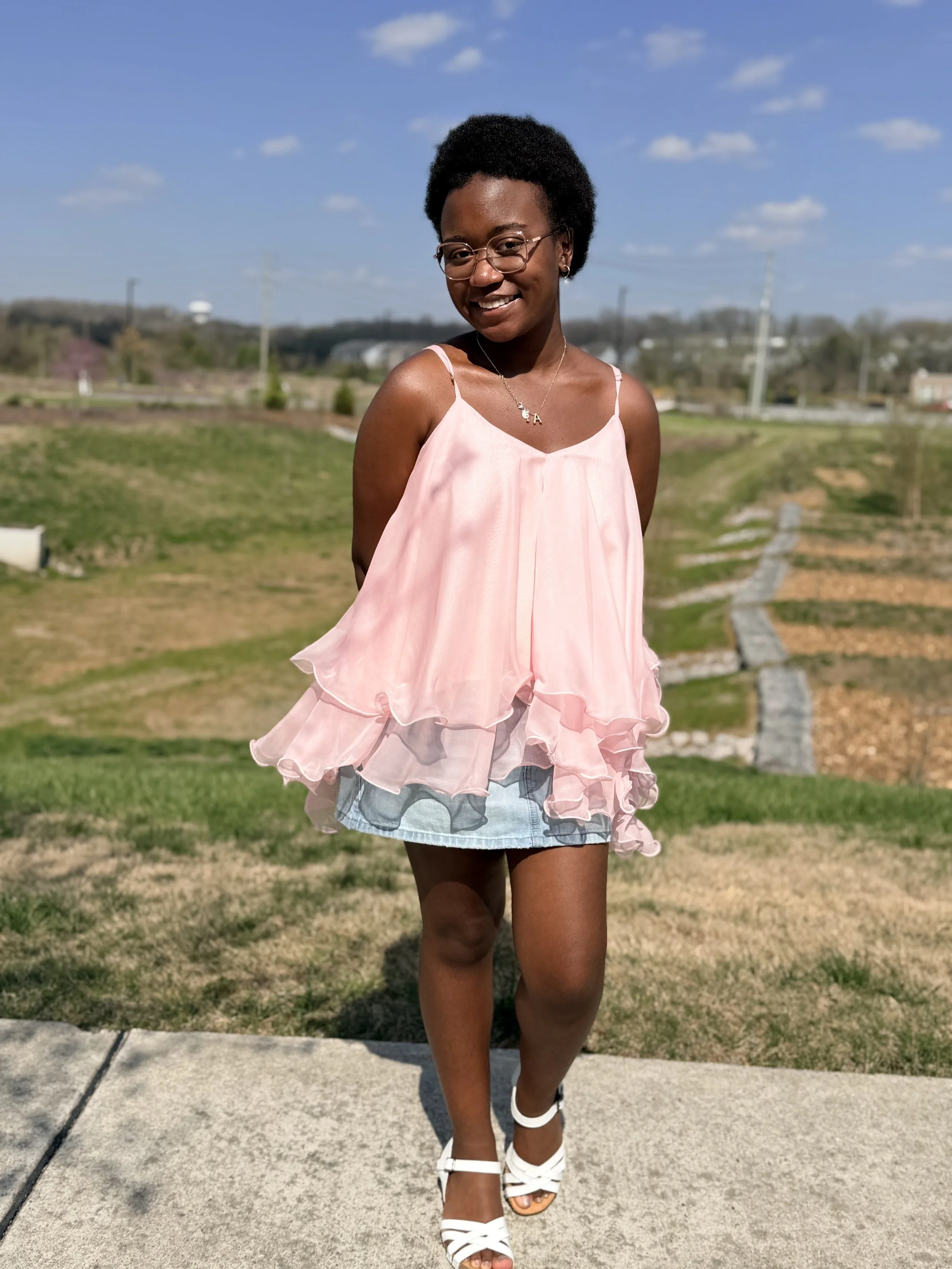 A young woman with short curly hair and glasses smiling while walking outdoors on a sunny day, wearing a light pink ruffled top, denim shorts, and white sandals. There is a grassy area, concrete sidewalk, and utility poles with a blue sky in the back