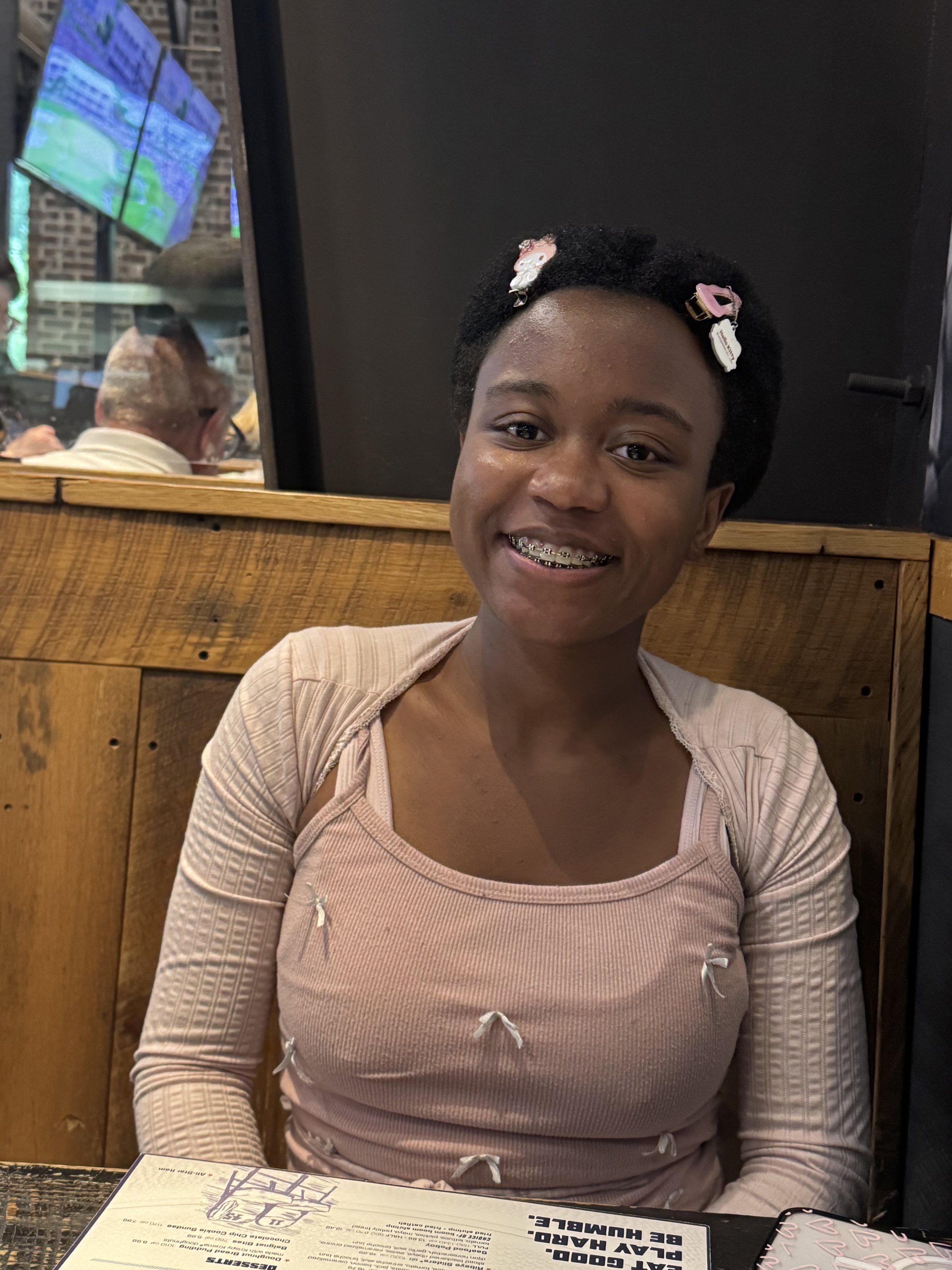 A smiling young girl with braces wearing a pink top and hair clips, sitting at a restaurant table with a menu in front of her.