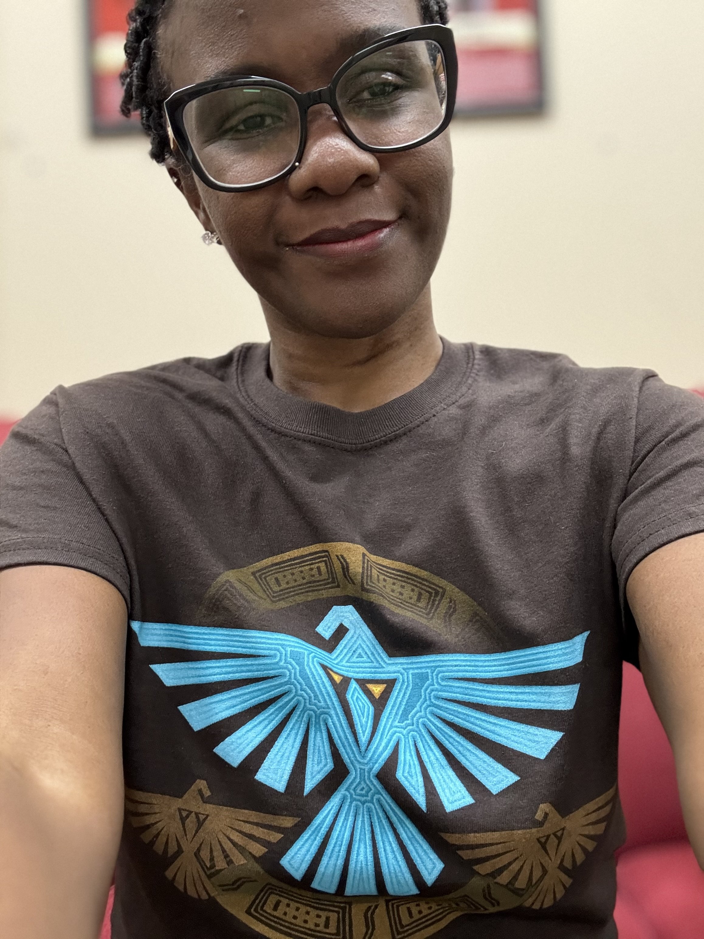 A woman taking a selfie, wearing glasses and a t-shirt with a distinctive blue and gold graphic of a bird with outstretched wings.