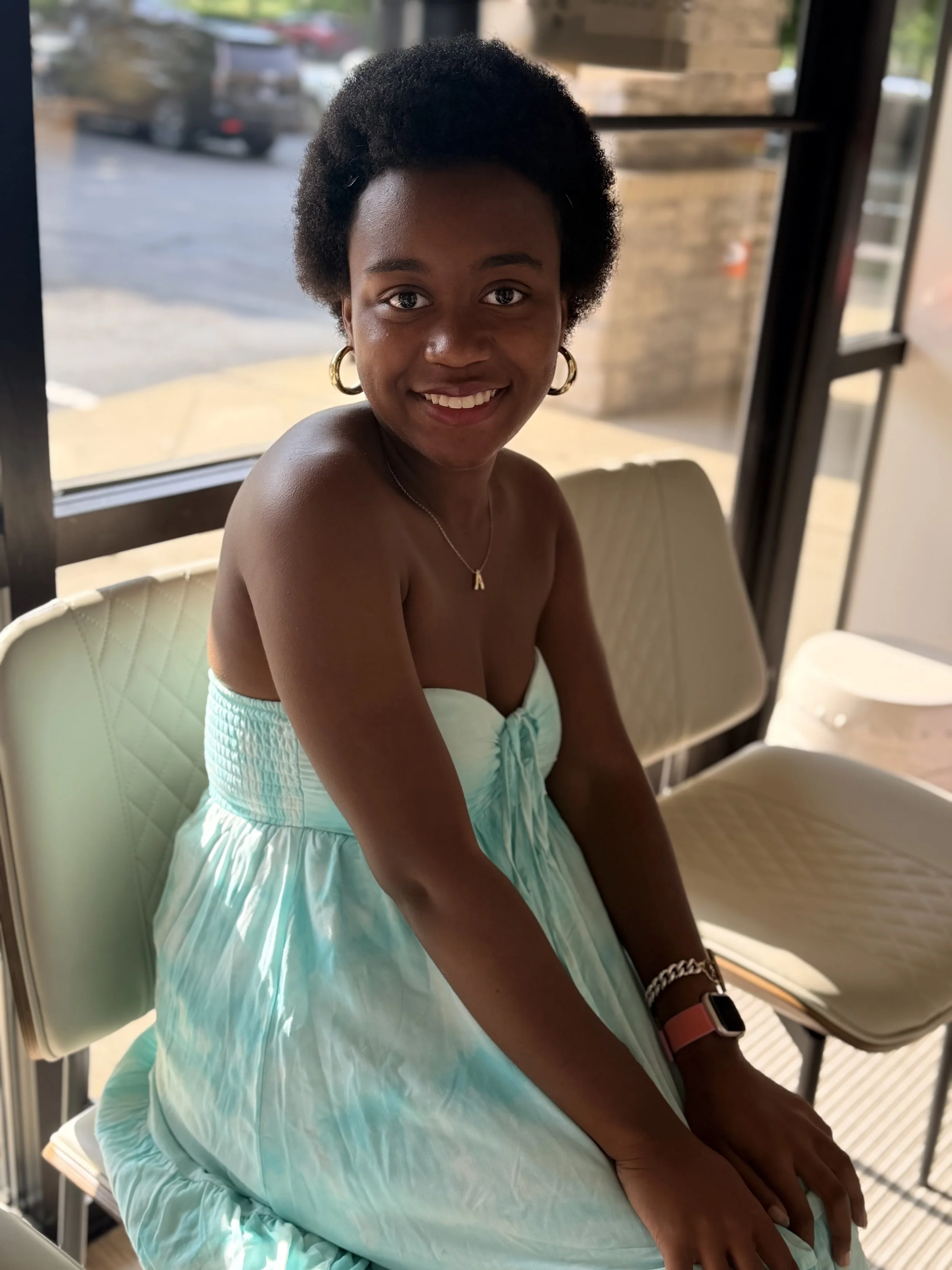 A young woman with short natural hair, hoop earrings, and a necklace with the letter 'A' sits on a cream-colored chair inside a building near a window, smiling at the camera.