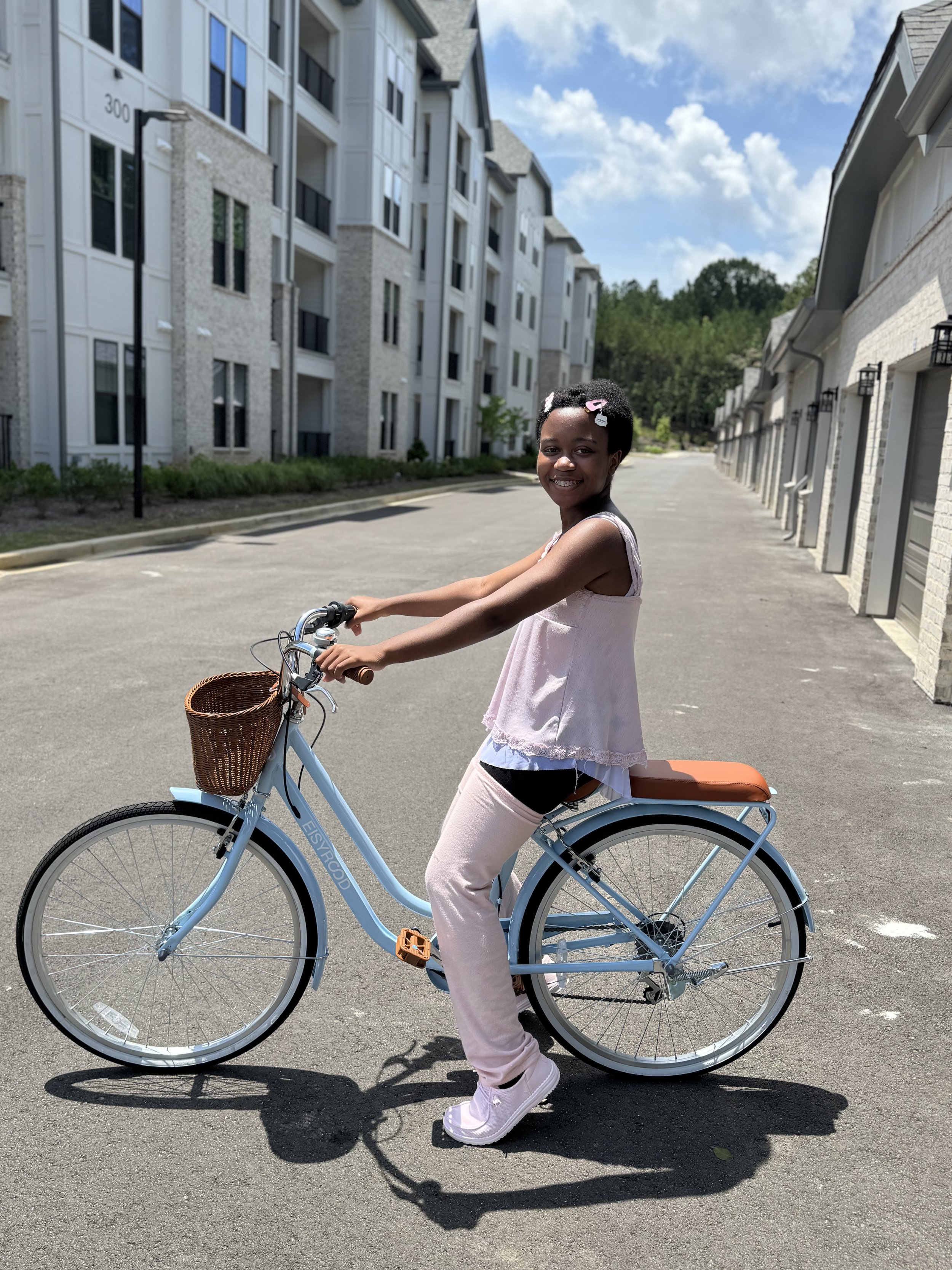 A smiling young girl on a light blue bike with a brown basket, riding on a paved path in front of modern white apartment buildings on a sunny day.