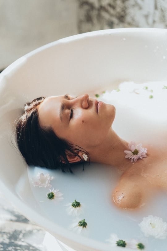 A woman lying back in a milky bath with a flower in her hair