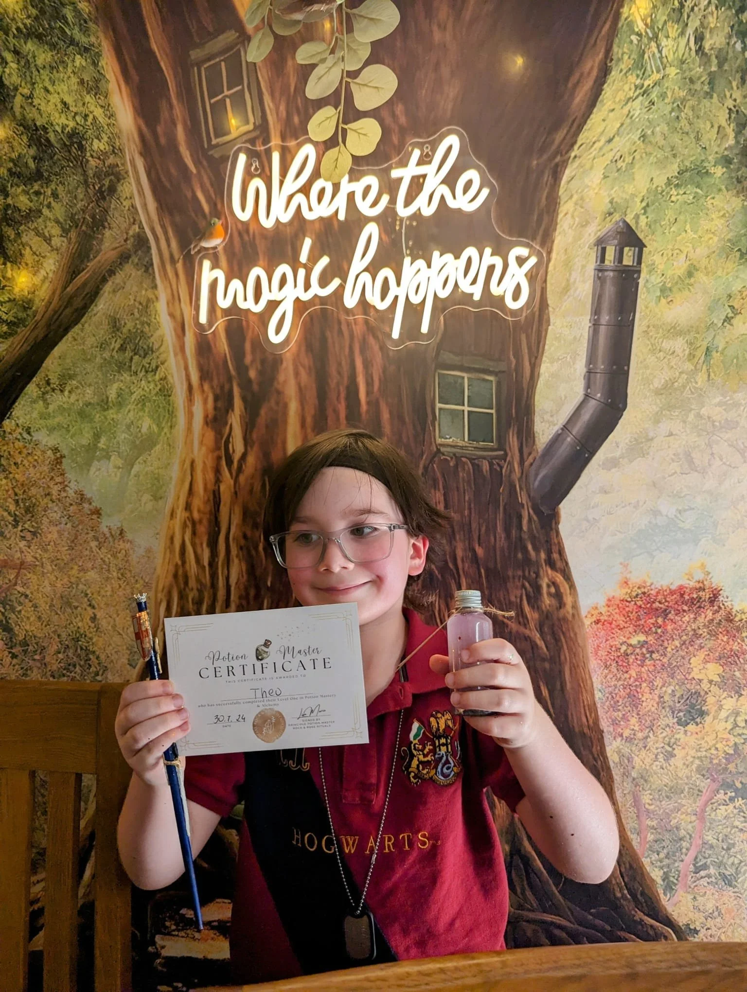 A young boy holding a potion master certificate and a small bottle, standing in front of a Harry Potter-themed backdrop with a large tree and the words 'where the magic happens'.