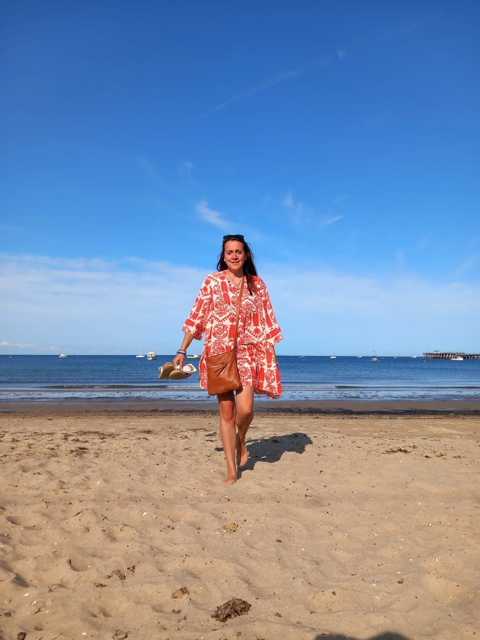 A woman walking barefoot on the sandy beach, holding flip-flops in one hand, with the ocean, boats, and clear blue sky in the background.