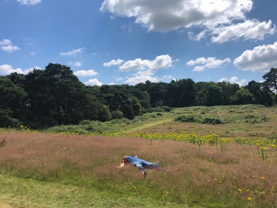 Person lying in a grassy field under a partly cloudy sky, with trees and hills in the background.