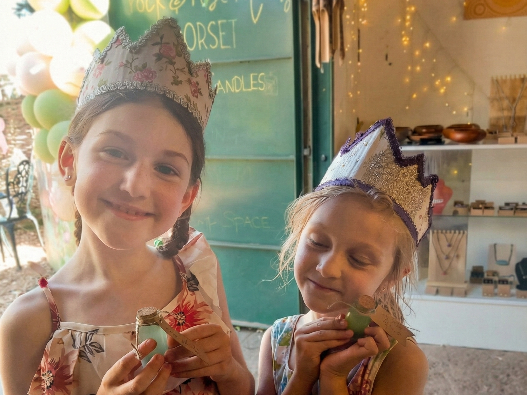 Two young girls wearing paper crowns with floral designs are smiling and holding small glass jars decorated with twine and tags. The girl on the left has braids and is smiling at the camera, while the girl on the right has curly hair and is looking down at her jar with a gentle smile. The background suggests they are at a celebration with balloons and fairy lights.