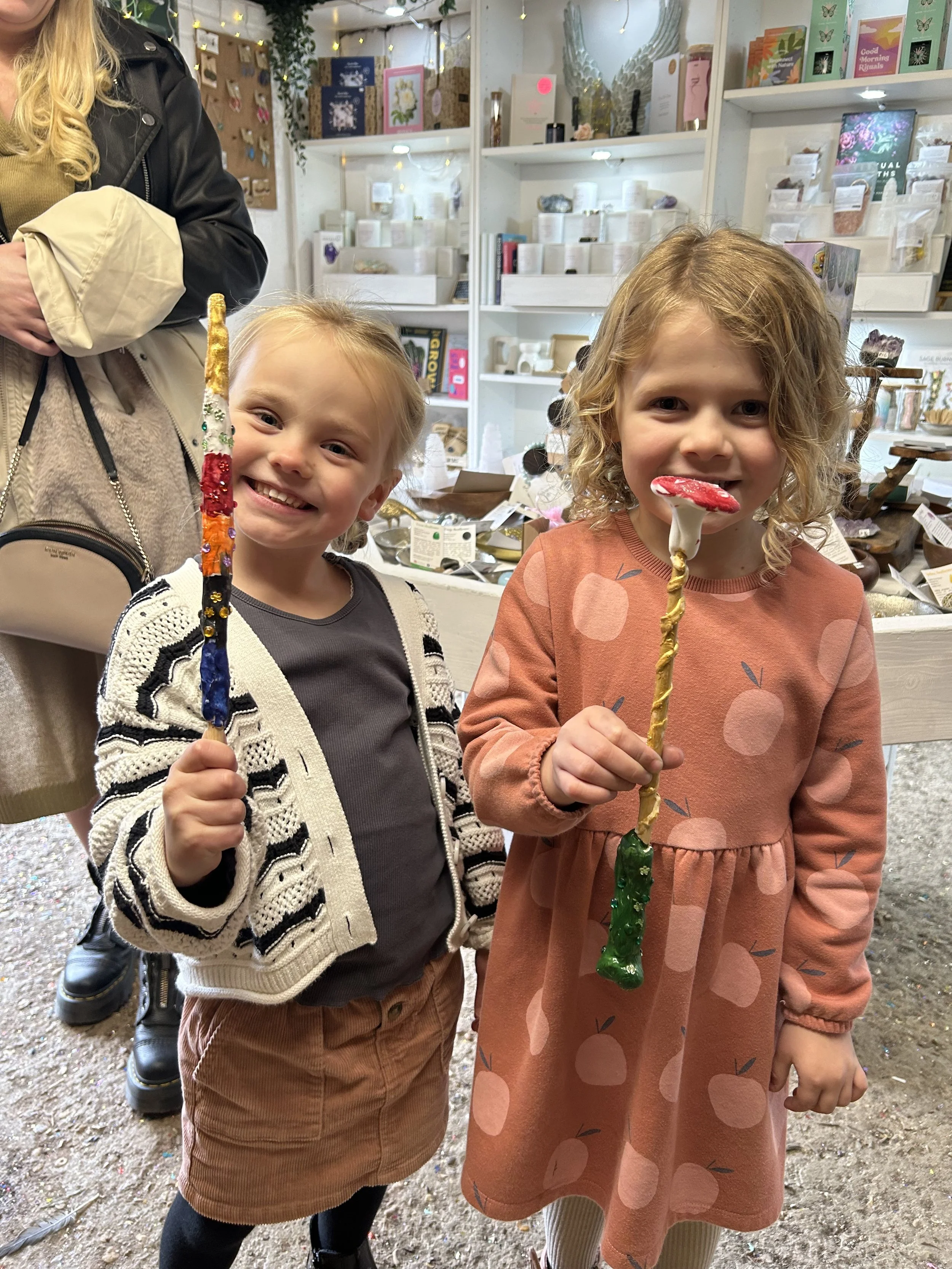 Two smiling young girls holding colorful candle-shaped lollipops inside a shop with shelves of crystals, jewelry, and gift items.