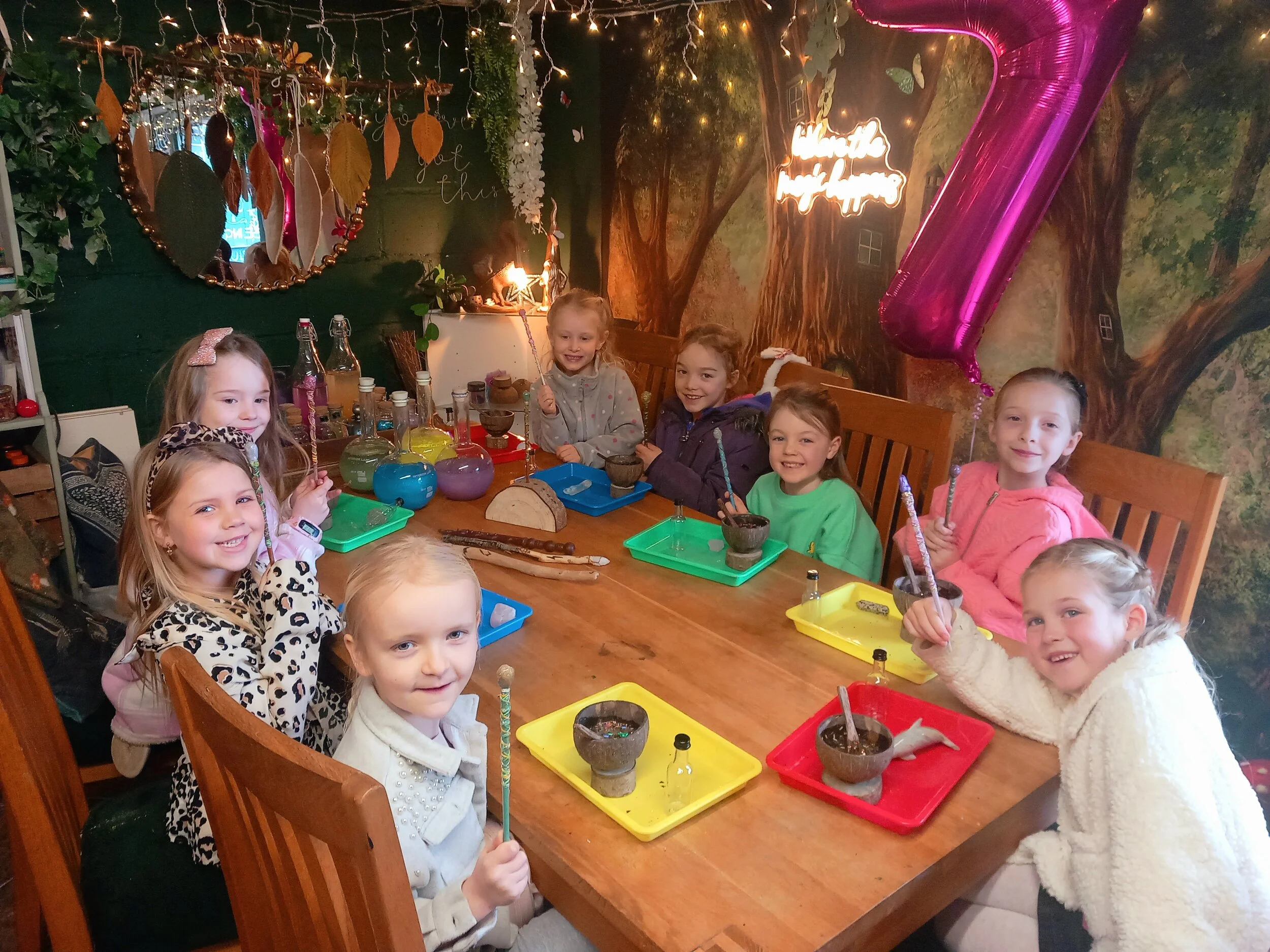 Group of young girls celebrating a birthday party at a decorated indoor setting with a large pink number 7 balloon, colorful trays, and a jungle themed wall with fairy lights.