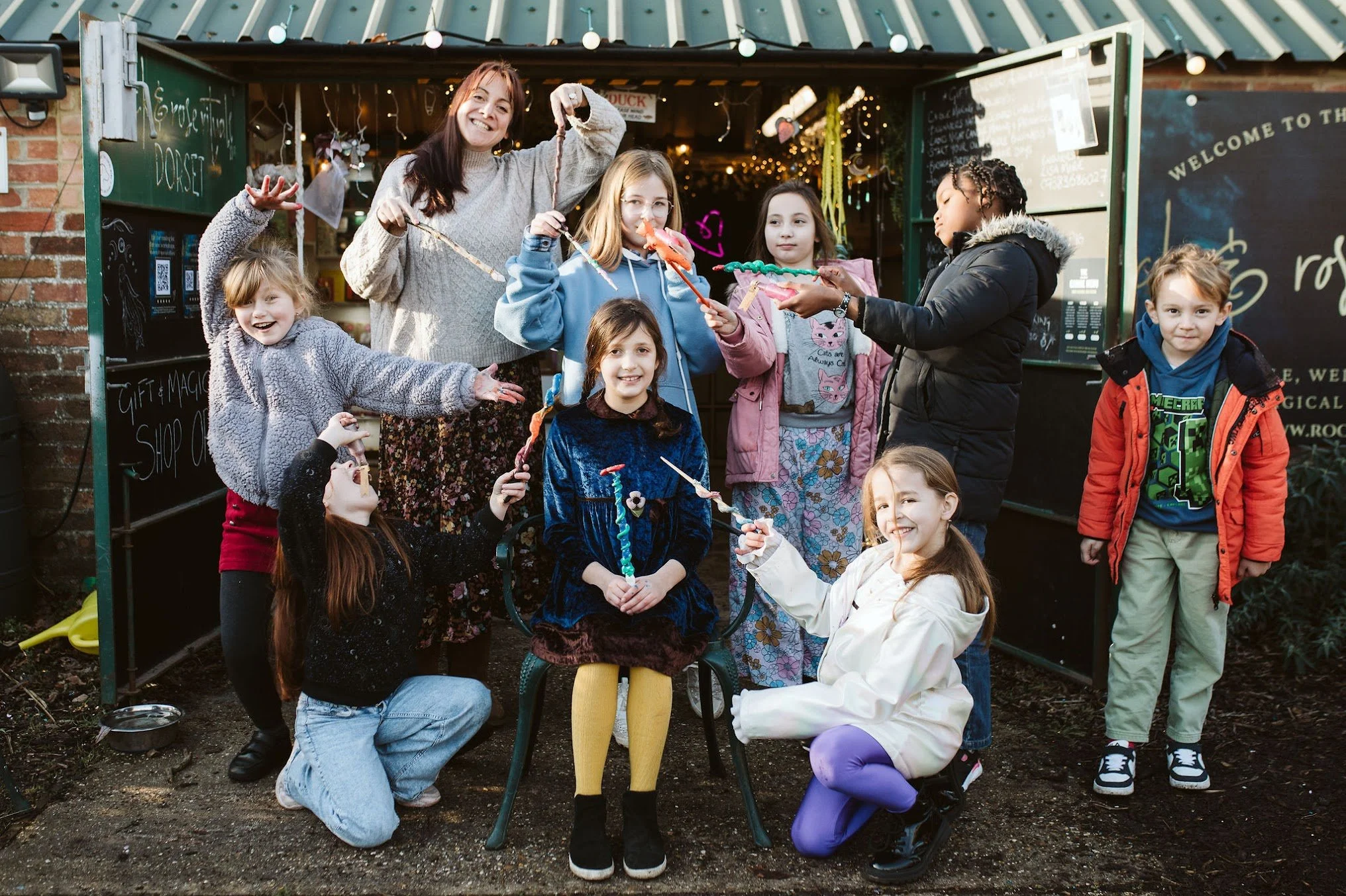 Group of children and a woman standing and sitting outside at a party, holding colorful candy skewers in front of a decorated storefront with string lights.