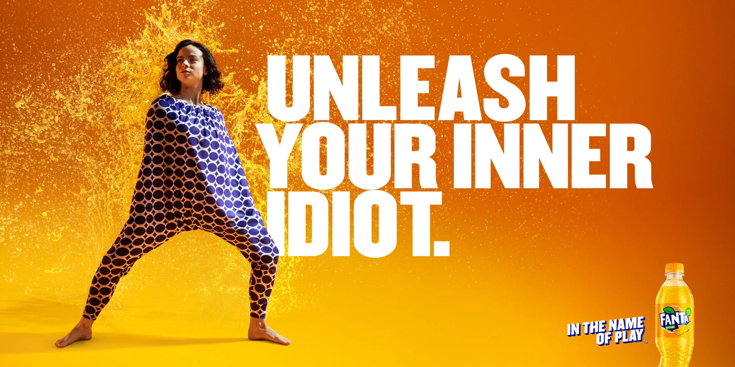A woman in dynamic pose wearing blue leggings with black circles, standing barefoot on a yellow surface with a vibrant orange background. Bold white text reads: 'Unleash Your Inner Idiot.' A small image of a Fanta soda bottle and the slogan 'In the Name of Play' are in the bottom right corner.