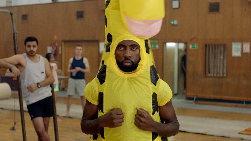 A person dressed in a Pikachu costume, standing in a gym or indoor sports facility, with other people in casual workout attire in the background.