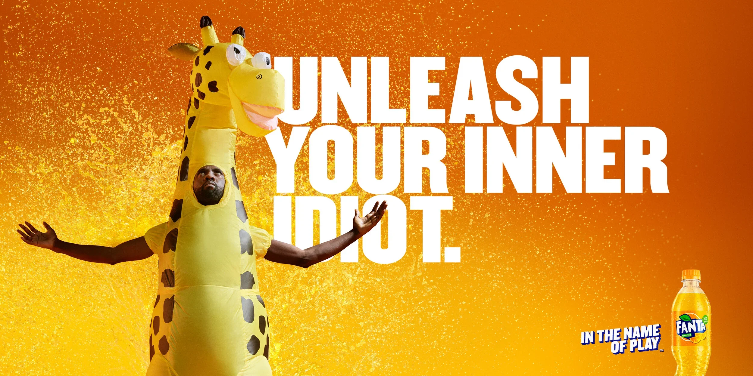 A person dressed in a giraffe costume with a human face emerging from the costume's neck, with arms outstretched, set against a bright orange background with yellow splashes. The text overlay says "Unleash Your Inner Idiot" and features a Fanta soda bottle with the slogan "In the Name of Play."
