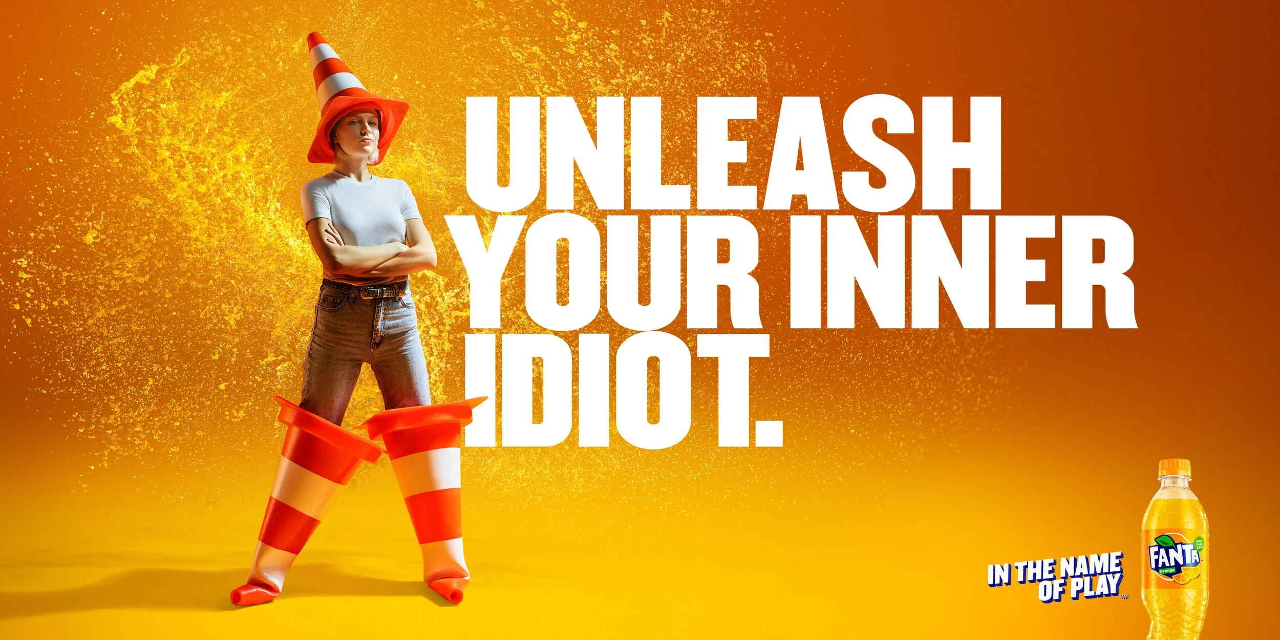 A woman stands with her arms crossed, wearing an orange and white striped traffic cone hat and matching oversized rain boots. The background is orange with yellow splatters, and there is bold white text that says "Unleash Your Inner Idiot." In the bottom right corner, there is a bottle of Fanta with the slogan "In the Name of Play."